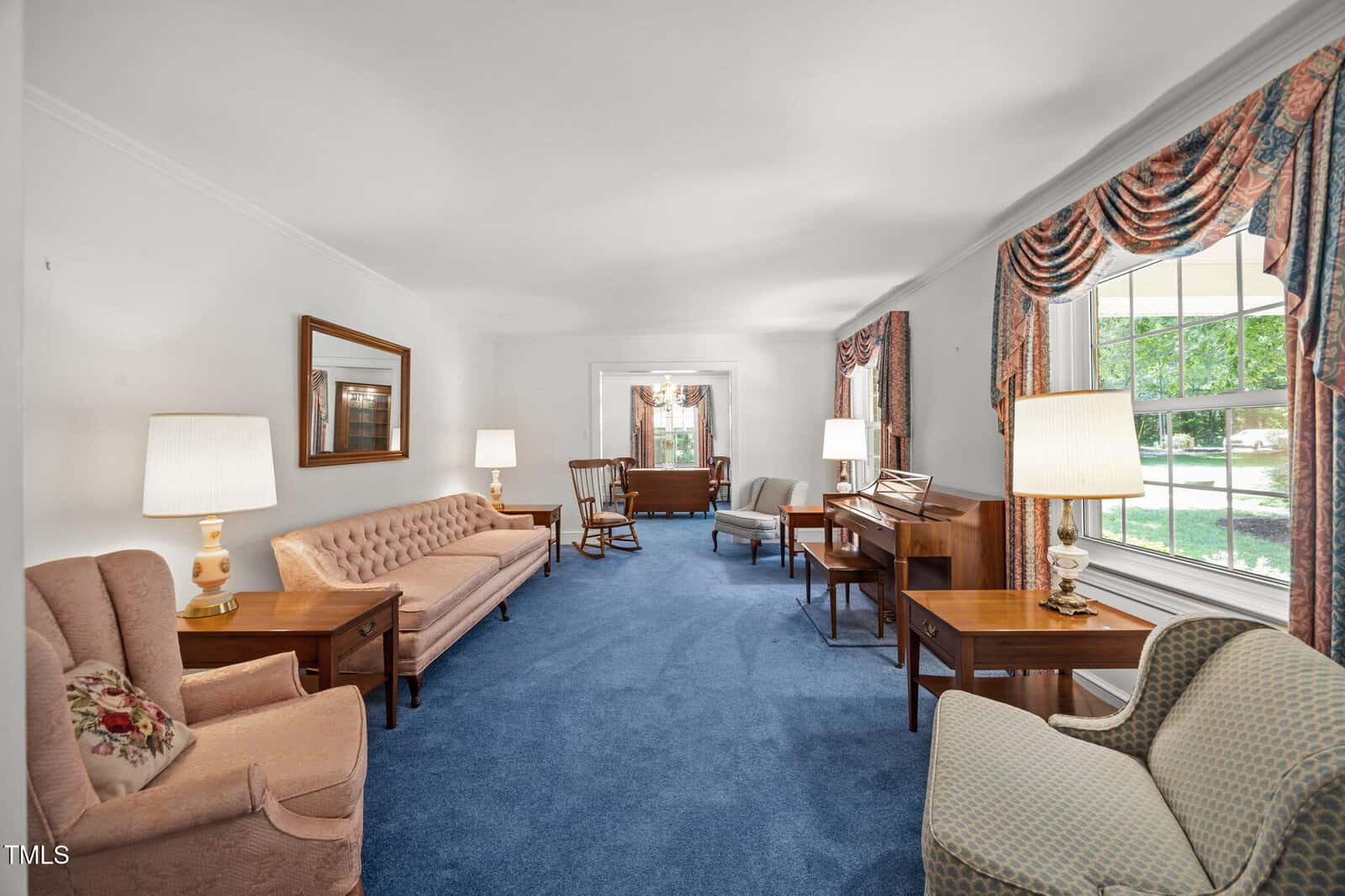 A traditional living room with blue carpet, pastel sofas and armchairs, wooden tables, lamps, and floral curtains framing a large window letting in natural light.