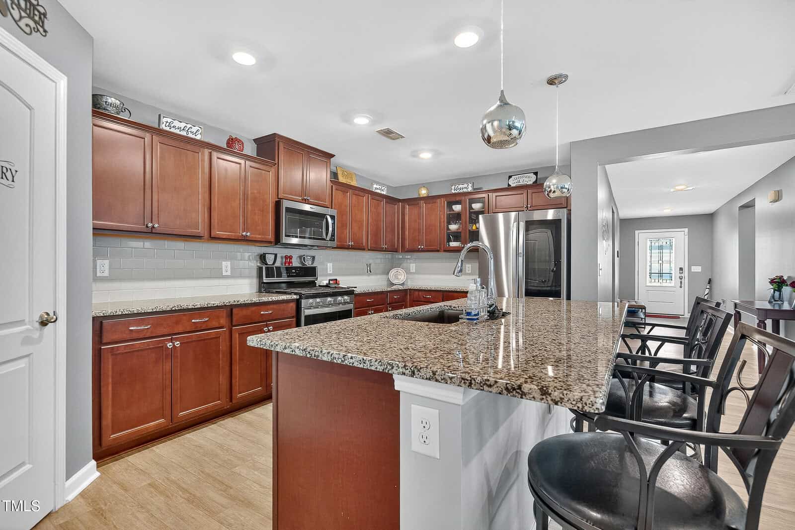 Modern kitchen with wooden cabinets, granite countertops, stainless steel appliances, and a large island with barstool seating. Pendant lights hang above the island; a hallway and a door are visible in the background.