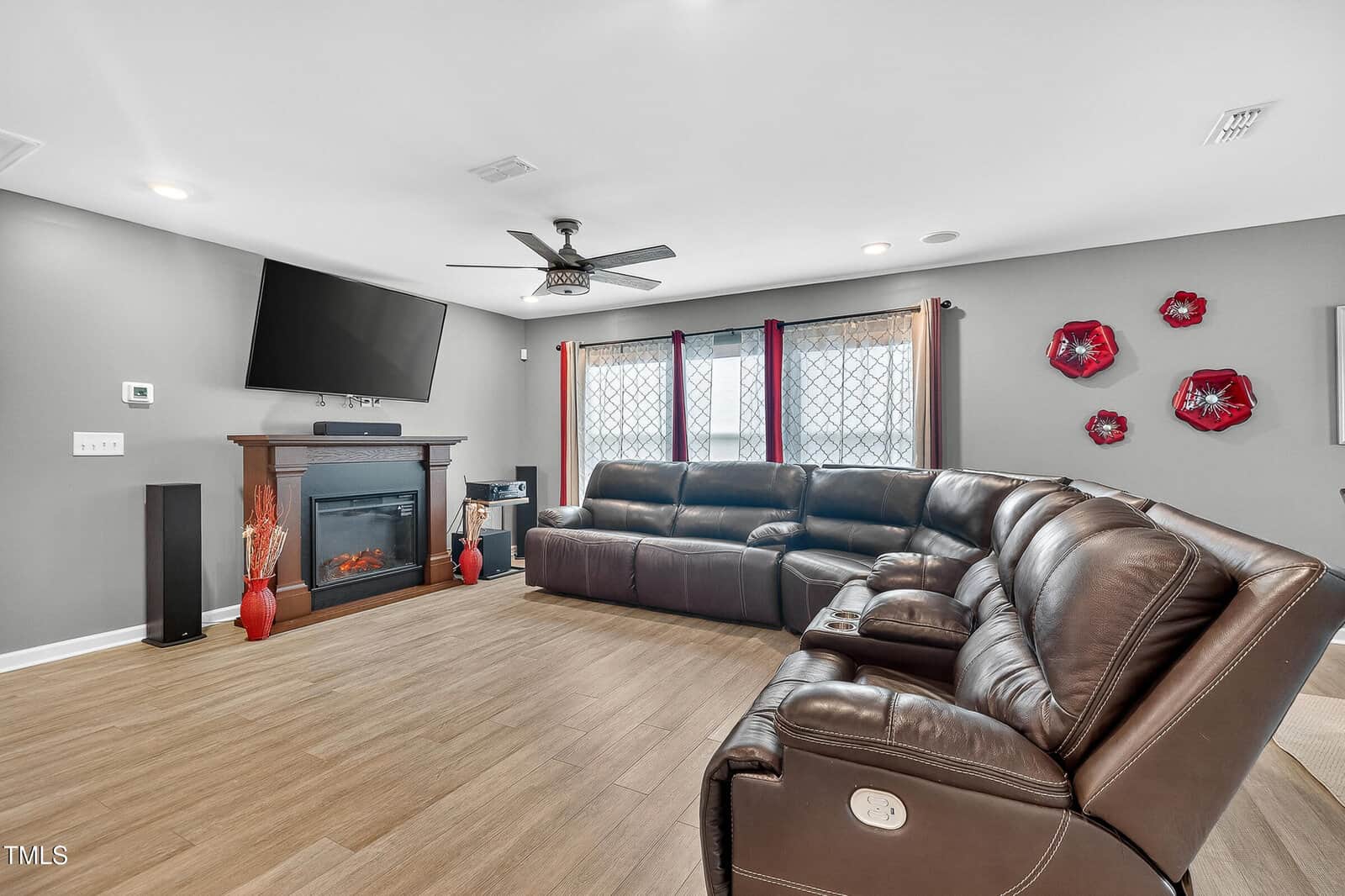 A modern living room with gray walls, wood flooring, a large brown sectional sofa, wall-mounted TV above an electric fireplace, and three red decorative plates on the wall.
