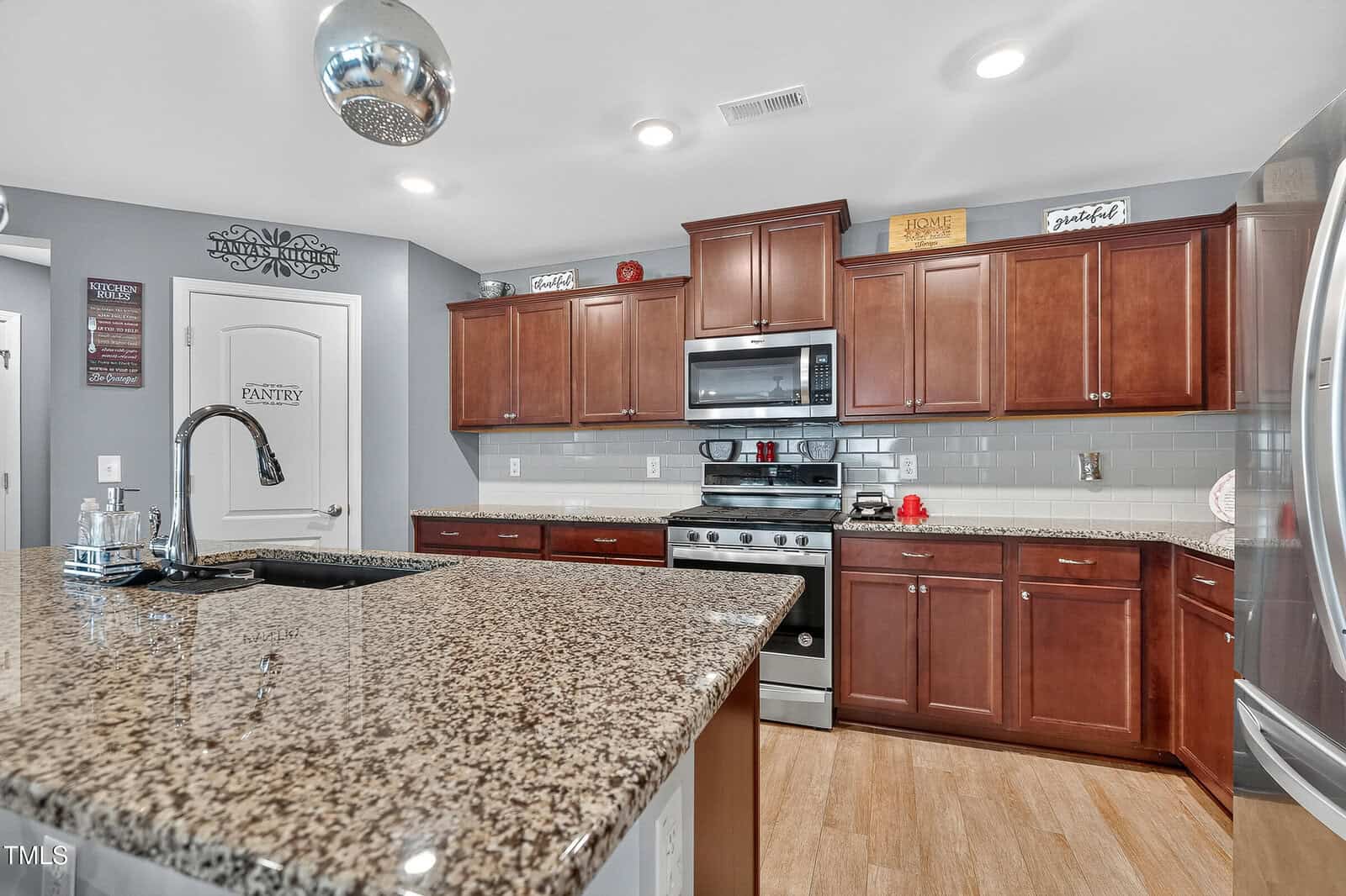 Modern kitchen with brown wood cabinets, stainless steel appliances, granite countertops, an island with a sink, and gray subway tile backsplash. Decorative signs and containers are displayed above the cabinets.
