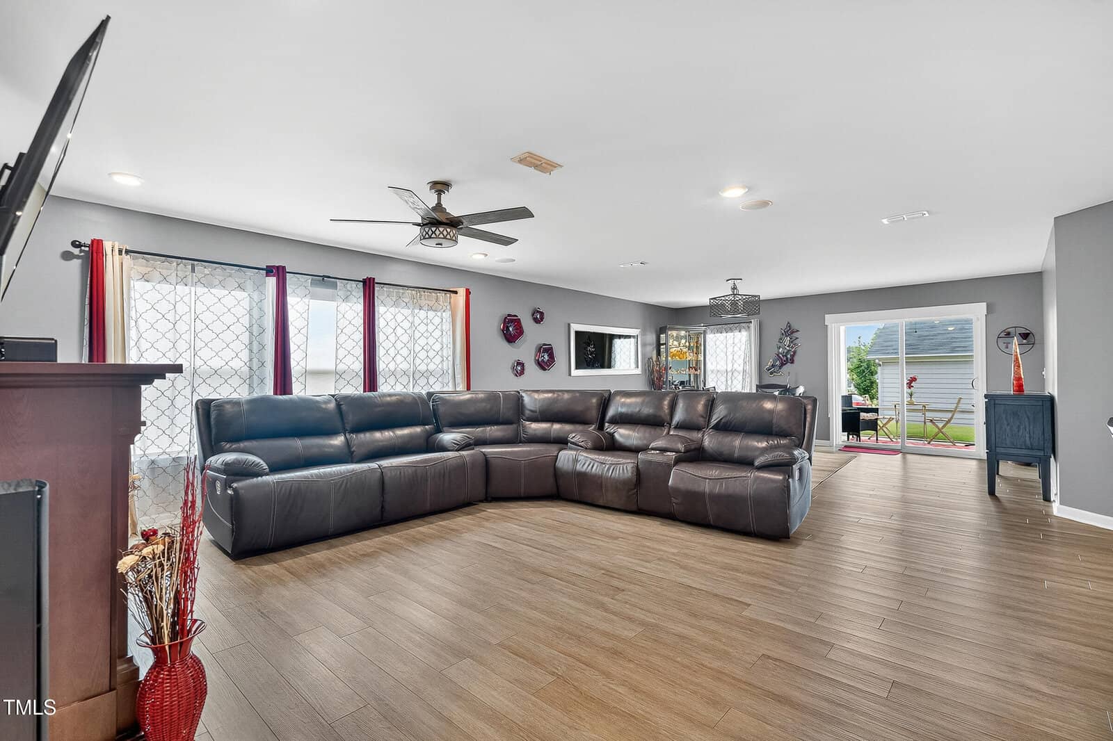A spacious living room with wood flooring, a large brown sectional sofa, wall-mounted TV, ceiling fans, and large windows with patterned curtains letting in natural light. A dining area and backyard are visible in the background.