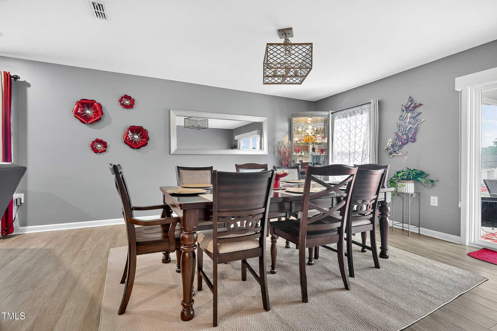A modern dining room with a dark wood table, six chairs, gray walls, red wall art, a large mirror, a china cabinet, butterfly wall decor, and light wood flooring. Natural light enters from a window and glass door.