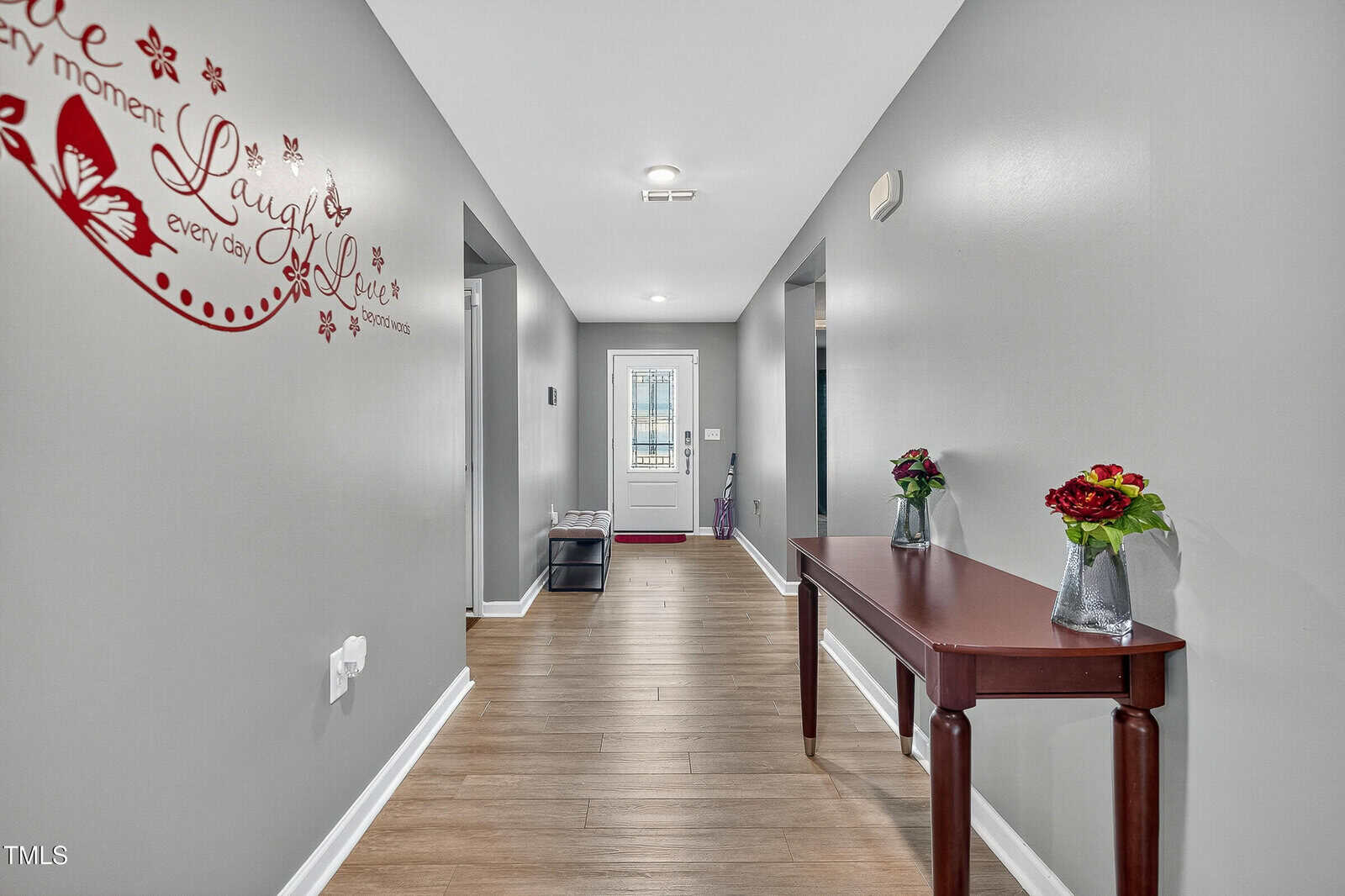 A hallway with gray walls, wood flooring, a red table with flower vases, wall art that says “Live, Laugh, Love,” and a door with a stained glass window at the end.