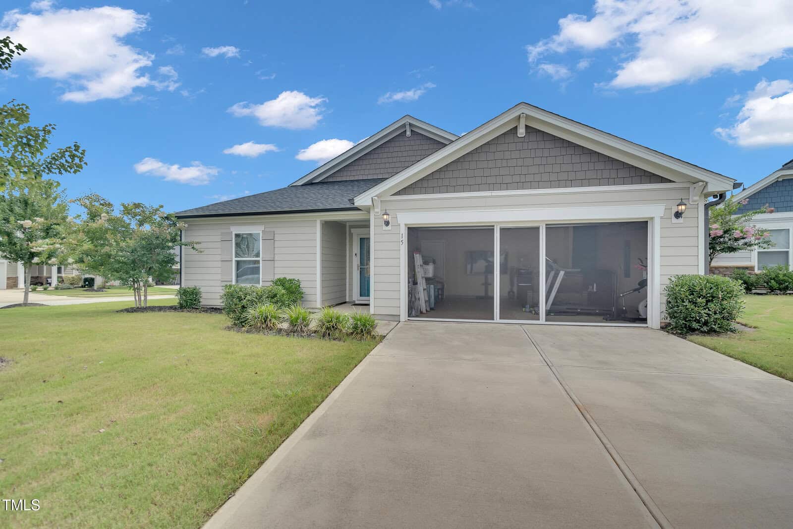 Single-story house with light gray siding, a gabled roof, and a two-car garage featuring a screened door. The house is surrounded by a green lawn, small shrubs, and bordered by a driveway under a blue sky with scattered clouds.