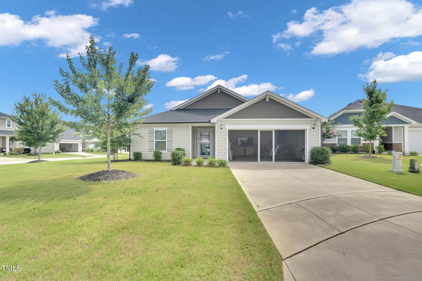 Single-story house with gray siding, a gabled roof, and a screened porch faces a wide driveway. The home is surrounded by a well-kept green lawn, small shrubs, and young trees under a blue sky with scattered clouds.