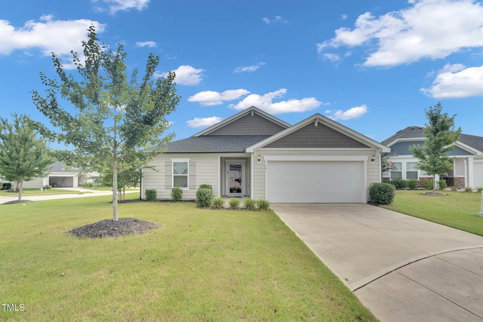 Single-story suburban house with gray siding, white trim, and a two-car garage. The front yard has green grass, small trees, and shrubs. The sky is blue with scattered clouds.