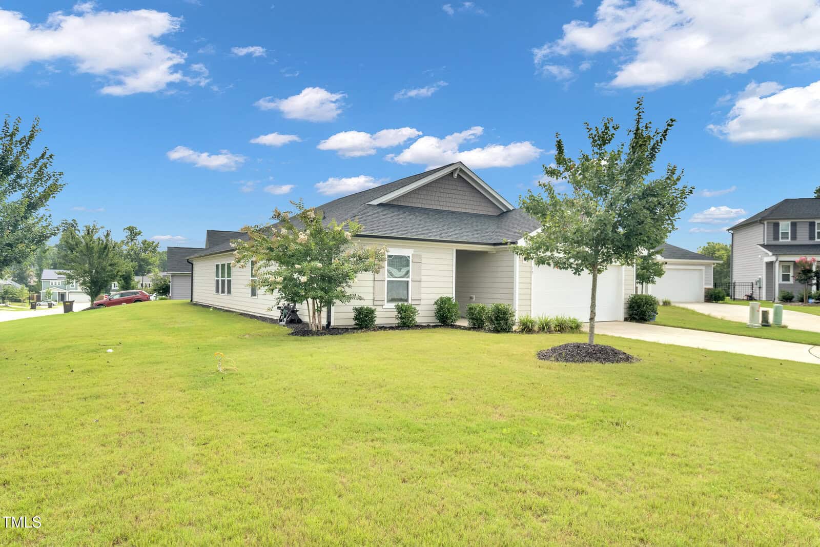A modern single-story house with gray siding and a dark roof sits on a large, well-manicured lawn with young trees. The sky above is blue with scattered clouds.