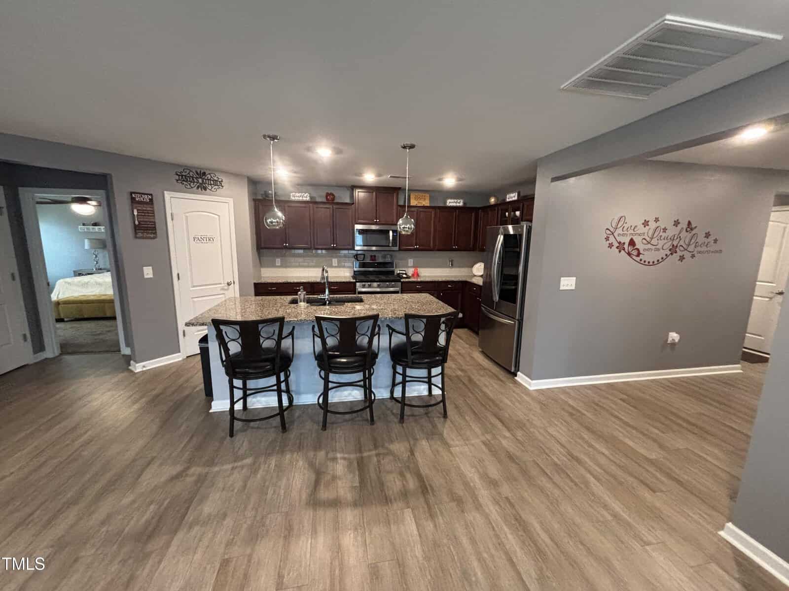 Modern kitchen with dark wood cabinets, stainless steel appliances, an island with granite countertop, and three black barstools. Light wood flooring and gray walls with decorative wall art and signs are visible.