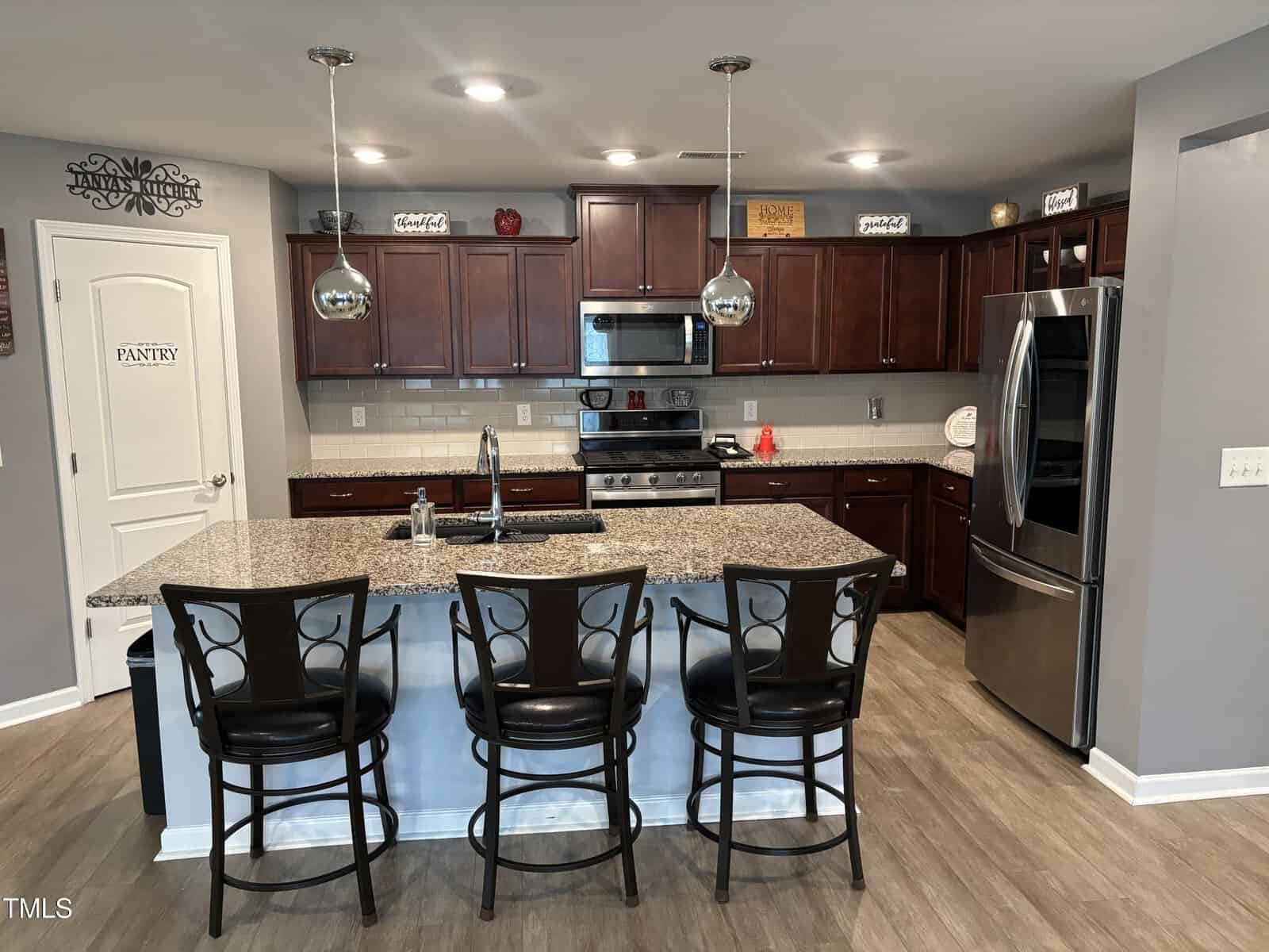 Modern kitchen with dark wood cabinets, stainless steel appliances, granite island with sink, three black chairs, and pendant lighting. Pantry door and decorative signs are visible above the cabinets.