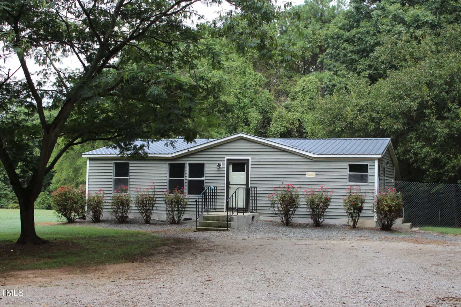 A single-story gray house with a metal roof at 350 Hamp Falkner Road, Henderson. Several windows and a small front porch with steps overlook bushes lining the front, while a gravel driveway leads to the entrance. Trees and greenery surround the property.
