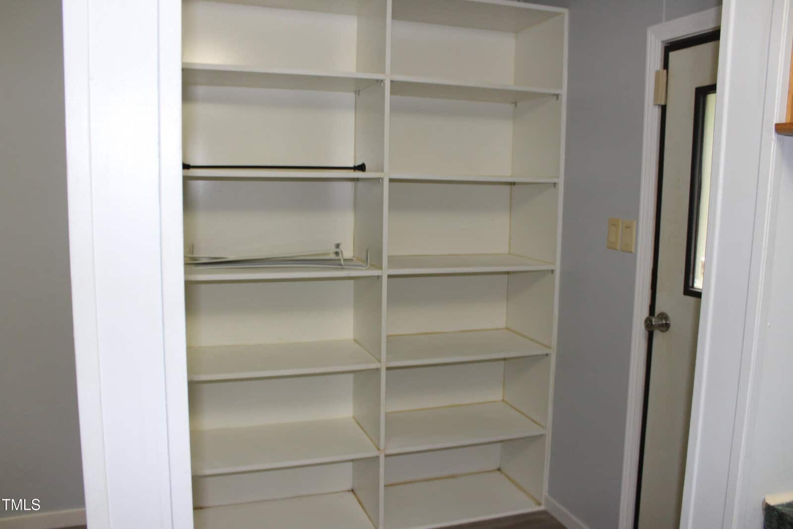 A built-in white shelving unit with eight shelves is installed against a light gray wall. An empty closet rod and some cords are visible on one shelf. A door with a window is on the right.