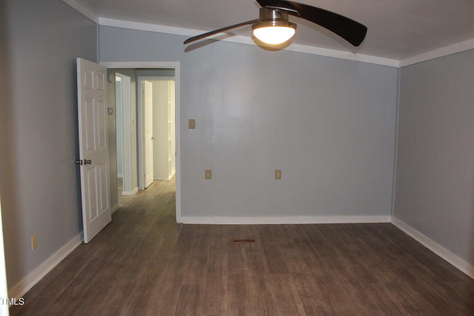 Empty room with gray walls, wood-look flooring, a ceiling fan with a light, and an open door leading to a hallway with additional doors visible.