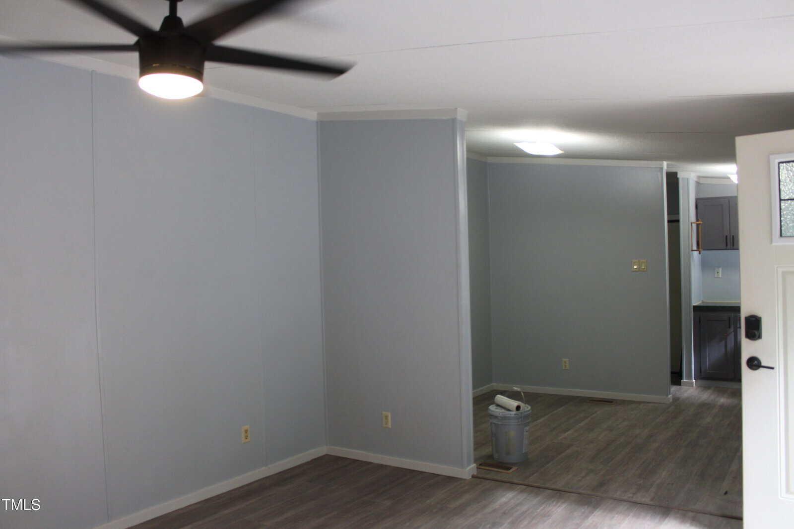 A room with light gray walls, a dark ceiling fan, and wood-style flooring. An adjacent area contains a paint bucket and supplies, and part of a kitchen is visible in the background.