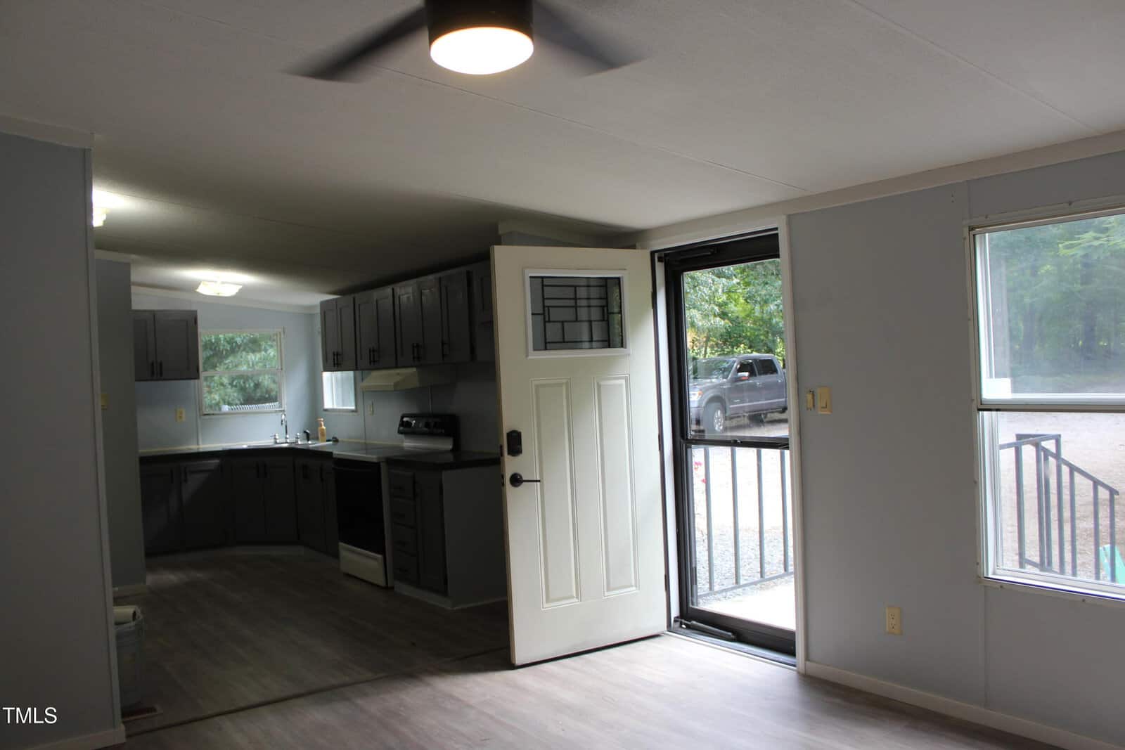 A view of a kitchen and entryway in a home with gray cabinets, light countertops, wood-style flooring, and an open door leading outside to steps and trees. A ceiling fan and large window are also visible.