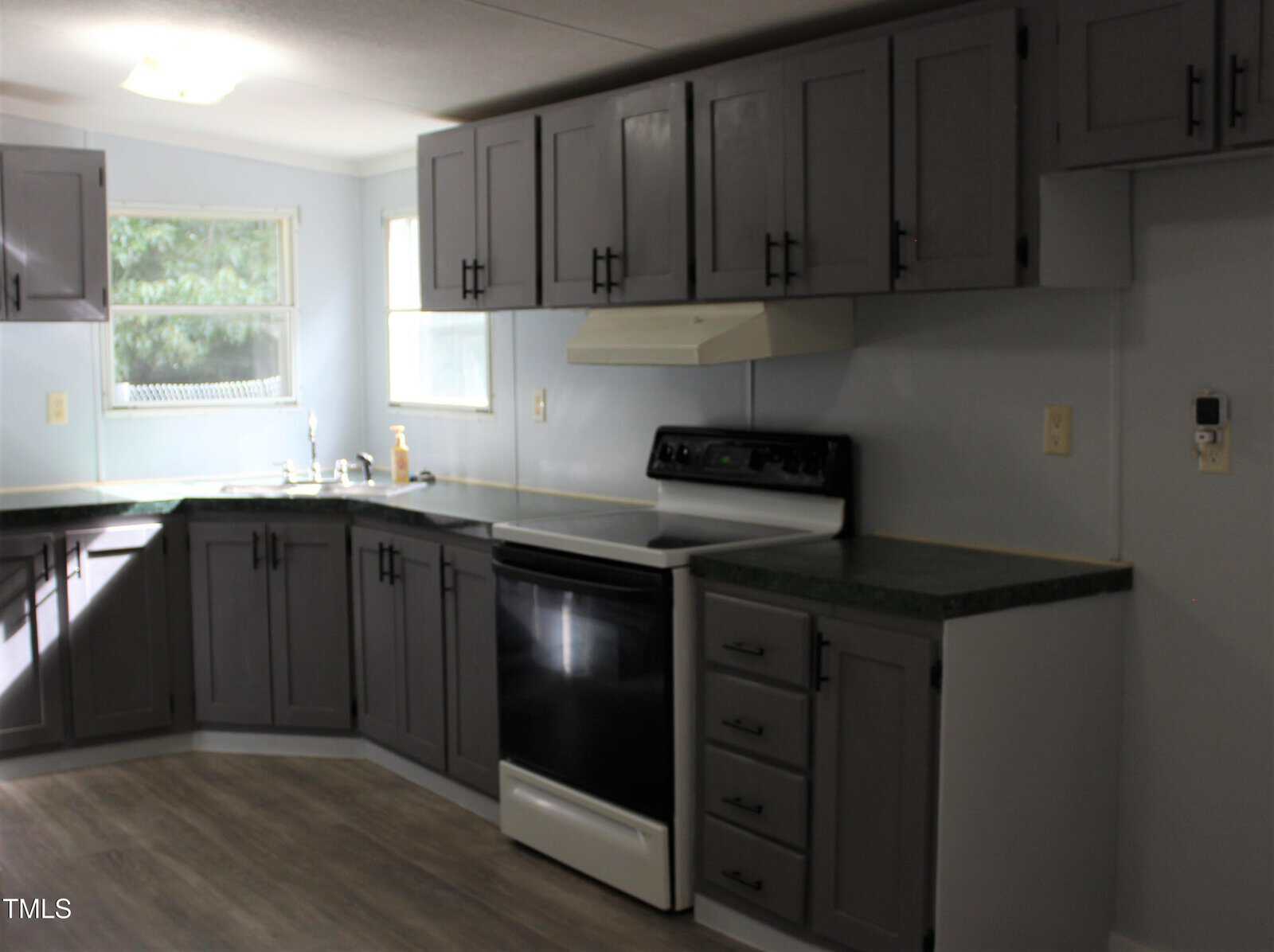 A kitchen with gray cabinets, black countertops, a white stove with a matching hood, a stainless steel sink under two windows, and light wood flooring. The walls are light blue, and there are no visible appliances on the counters.