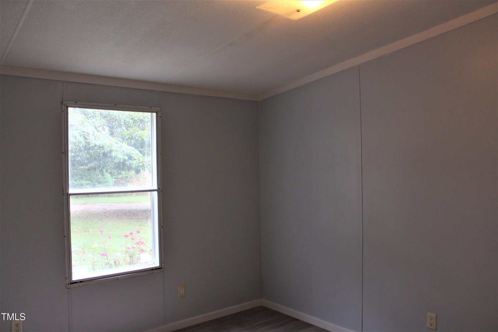 A small, empty room with light blue walls, a single window showing greenery outside, a white ceiling with a ceiling light, and gray flooring. There are two electrical outlets visible near the window.