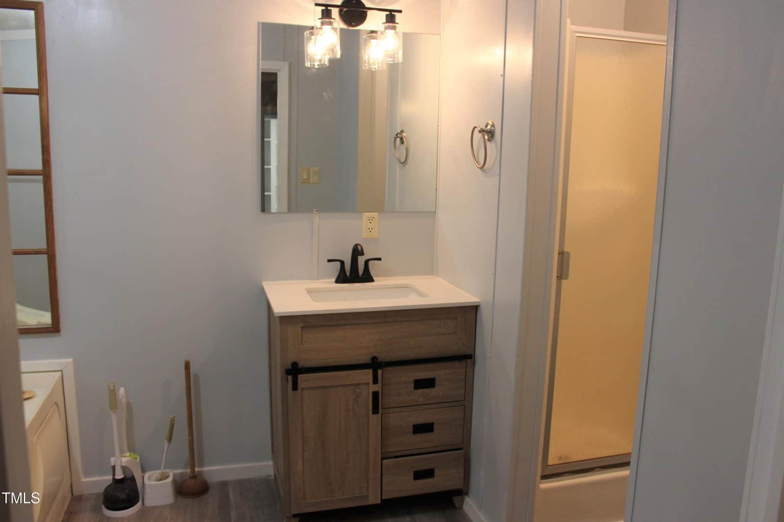 A bathroom features a vanity with a rectangular mirror, black faucet, and wooden cabinet, next to a shower with a frosted glass door. Cleaning tools, including a plunger and toilet brush, are on the left.