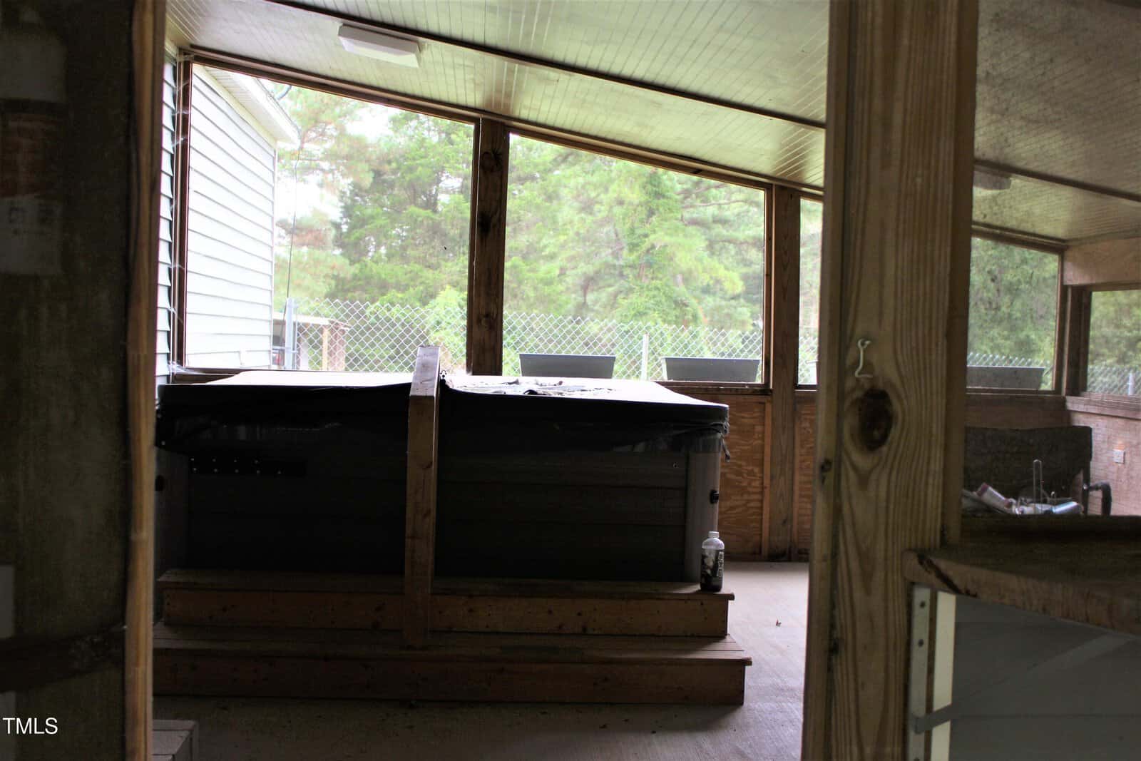 Indoor screened porch with a covered hot tub on a wooden platform, surrounded by wooden framing. There are windows looking out to a green yard with trees and a chain-link fence in the background.