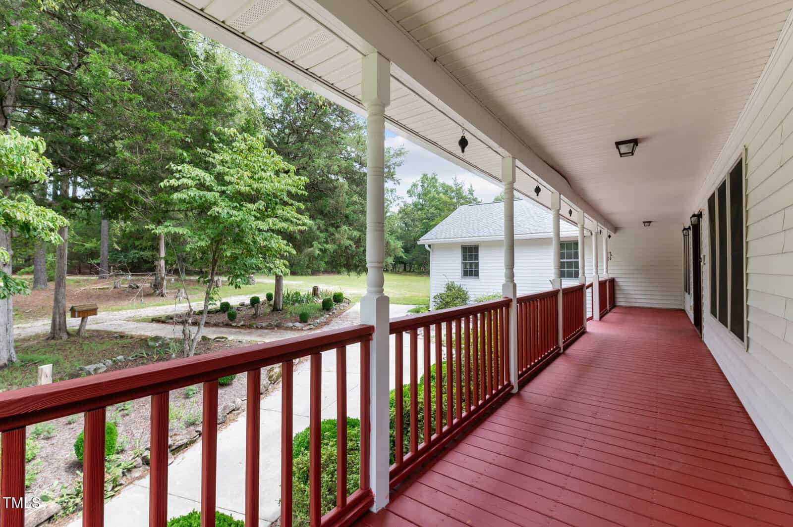 A long, covered front porch with red flooring and white railings overlooks a landscaped yard with trees and a detached white garage in the background.