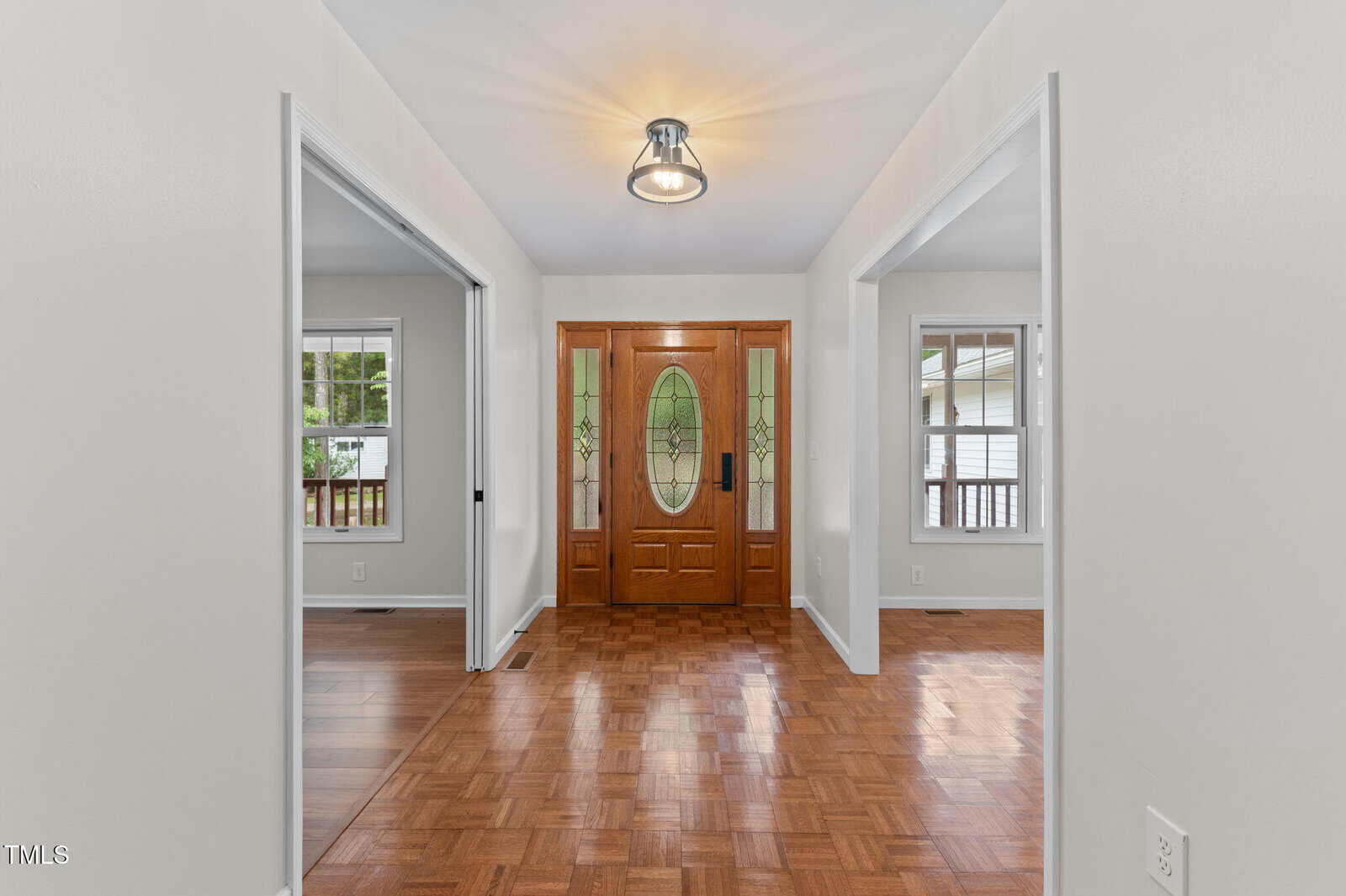 A wooden front door with decorative glass panels is centered in a white hallway with parquet flooring. Two rooms with windows are visible on either side, separated by open doorways. A ceiling light fixture hangs above.