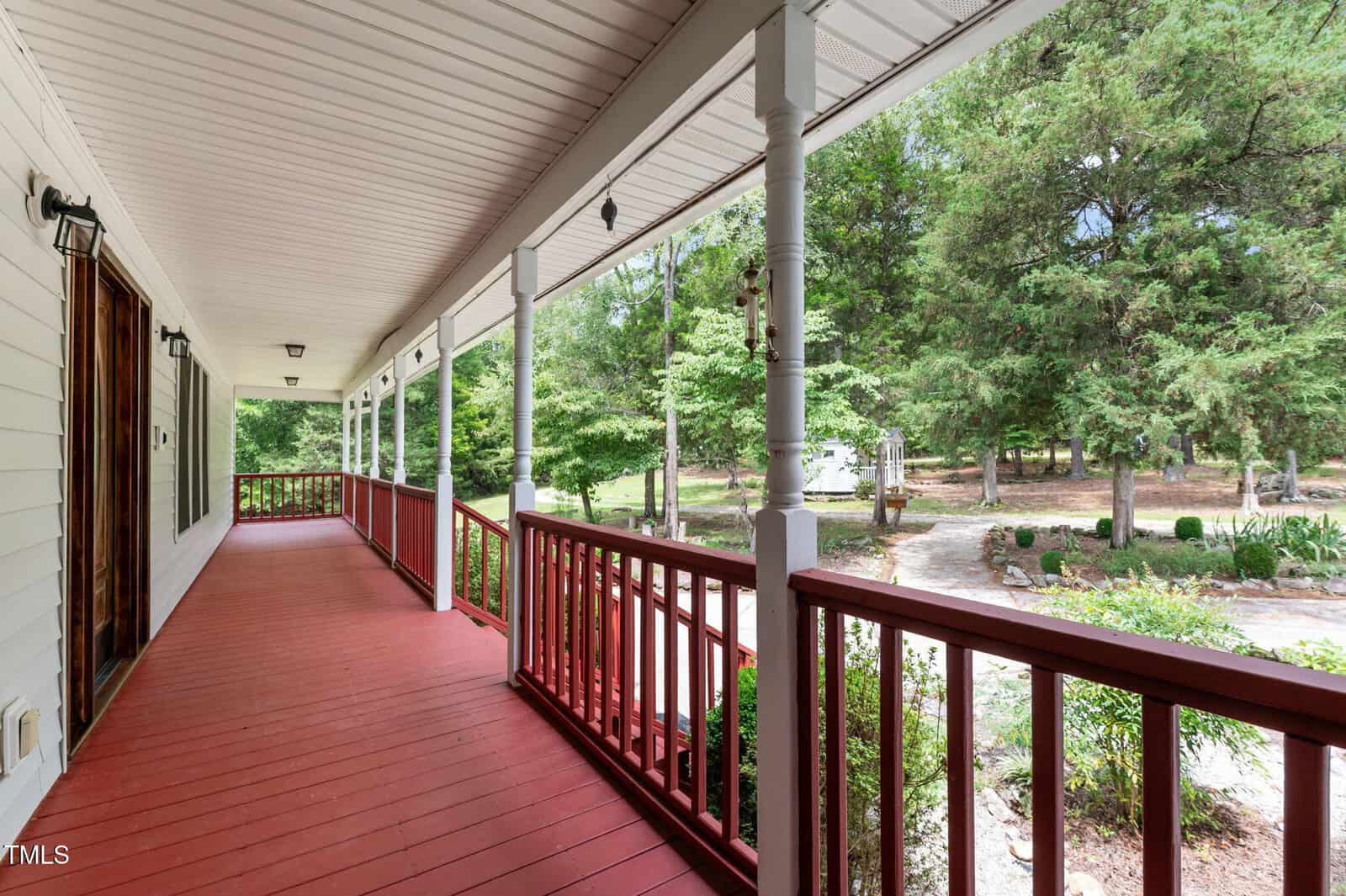 A covered porch with red flooring and railings stretches alongside a white house, overlooking a landscaped yard with trees, shrubs, a path, and garden beds under a cloudy sky.