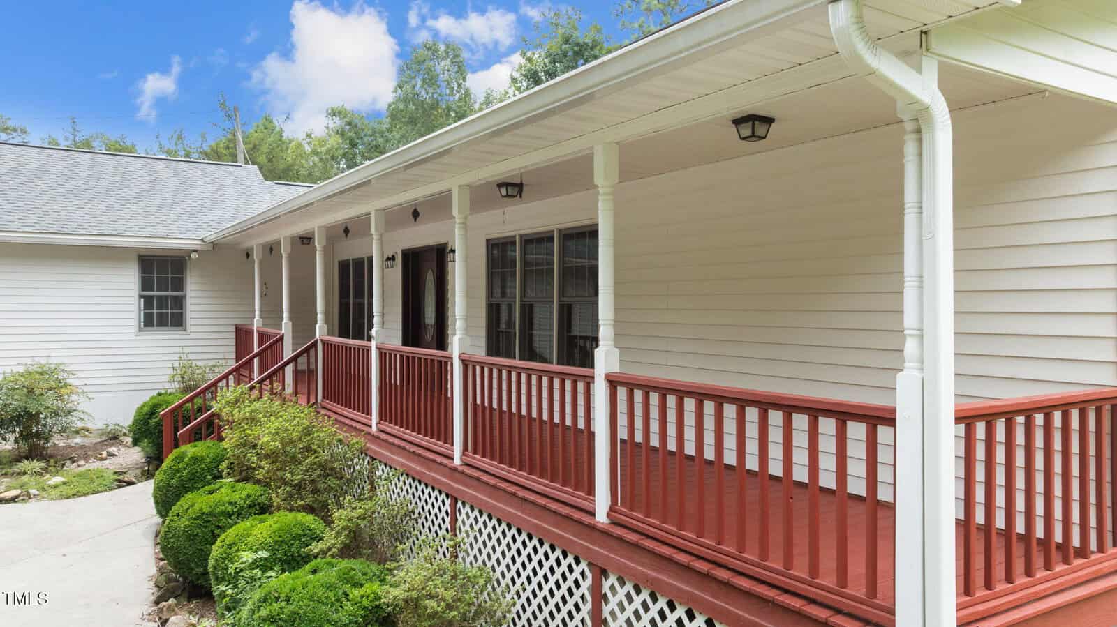 A white house with a covered front porch featuring red railings and posts. Shrubs line the walkway leading to the porch, and the sky is partly cloudy with trees in the background.