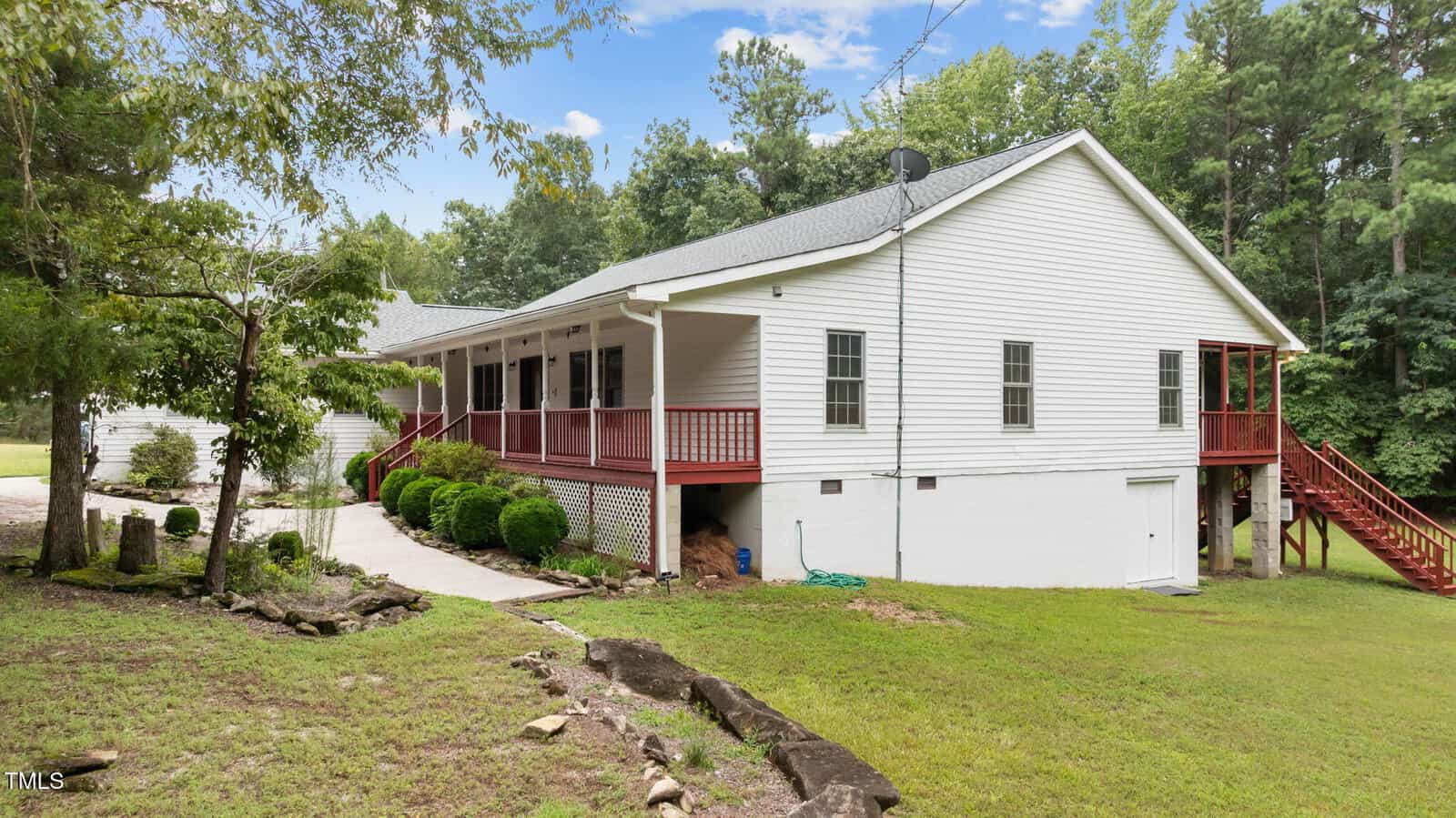 A white single-story house with a covered front porch, red railings, and a basement entrance. There are steps leading to a side door and green landscaping with bushes and trees surrounding the home.