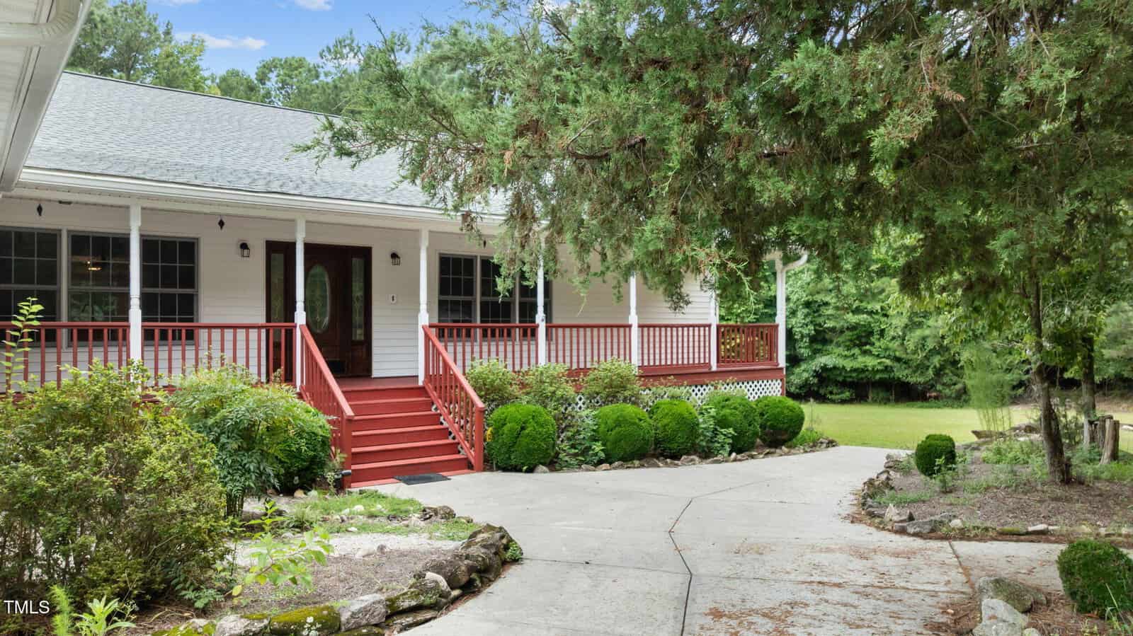 A white house with a wide front porch, red railings, and red steps, surrounded by green bushes and trees, with a curved concrete walkway leading to the entrance.