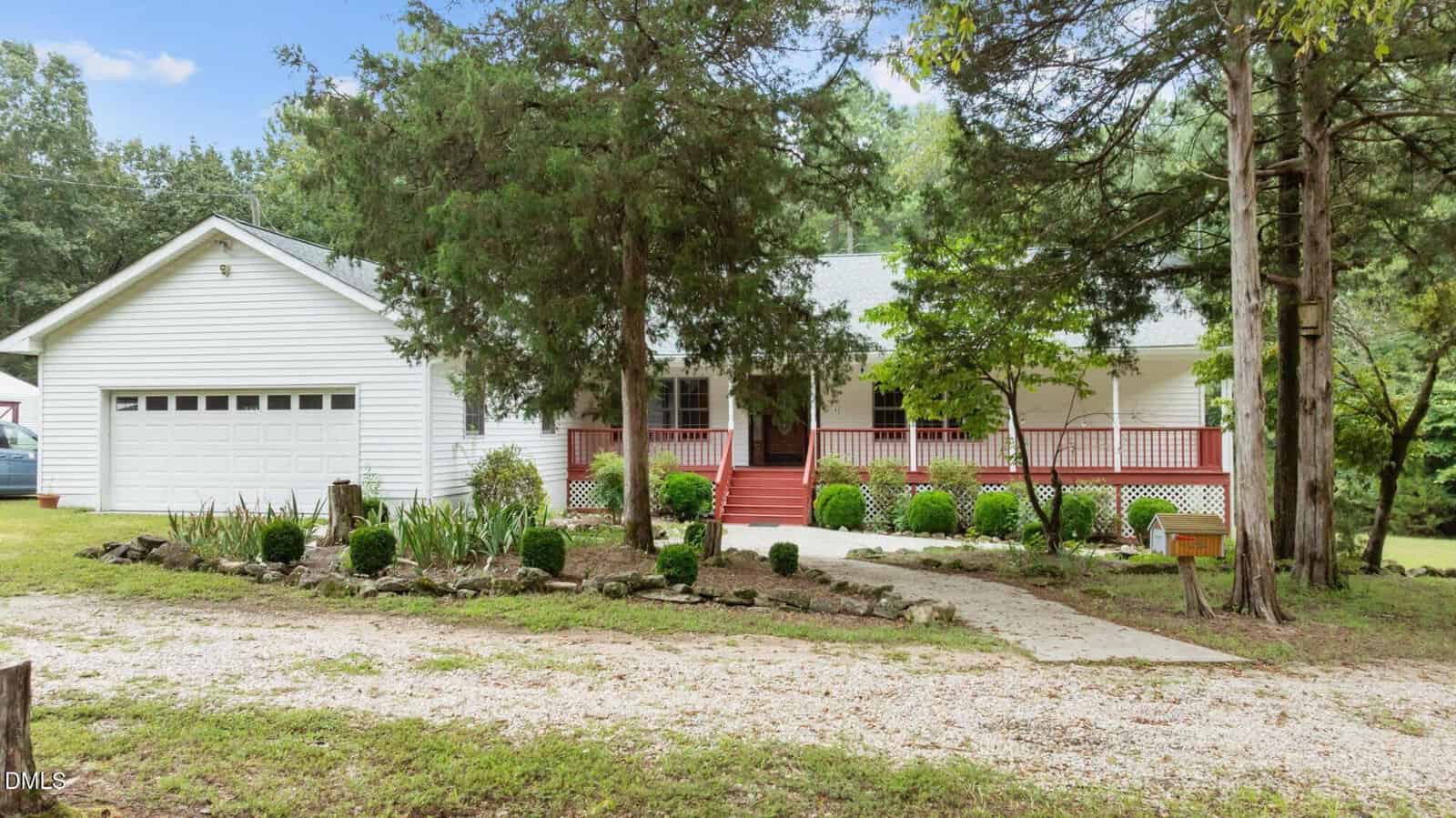 A white, single-story house with a front porch, red steps, and a two-car garage is surrounded by trees and greenery. A gravel driveway leads to the house, and landscaping lines the walkway to the entrance.