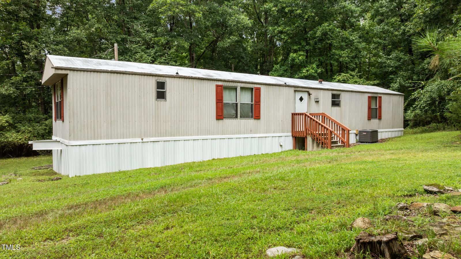 A beige mobile home with red shutters and a small wooden staircase sits on a grassy lot surrounded by trees and greenery.