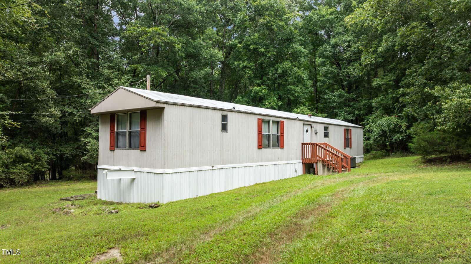 A beige single-wide mobile home with red shutters and a small front porch sits on a grassy lot surrounded by trees and forest.