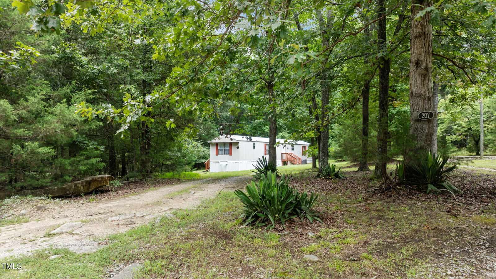 A white mobile home with a green roof sits among tall trees, at the end of a gravel driveway in a wooded area. The yard is shaded, with plants and rocks near the driveway and a house number sign on a tree.