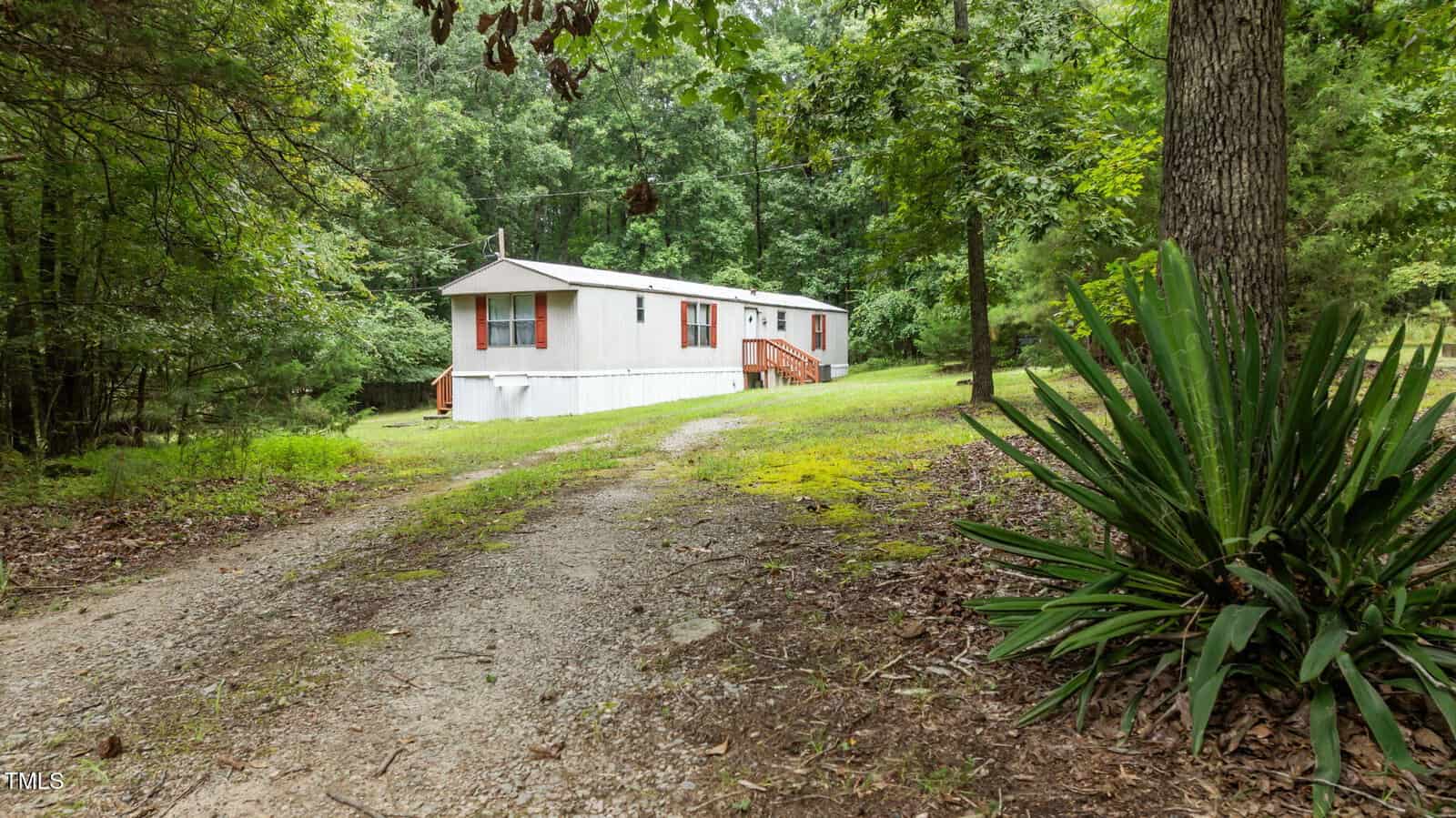 A white mobile home with red shutters sits surrounded by green trees and grass. A dirt driveway curves toward the home, and large leafy plants and tall trees are in the foreground.