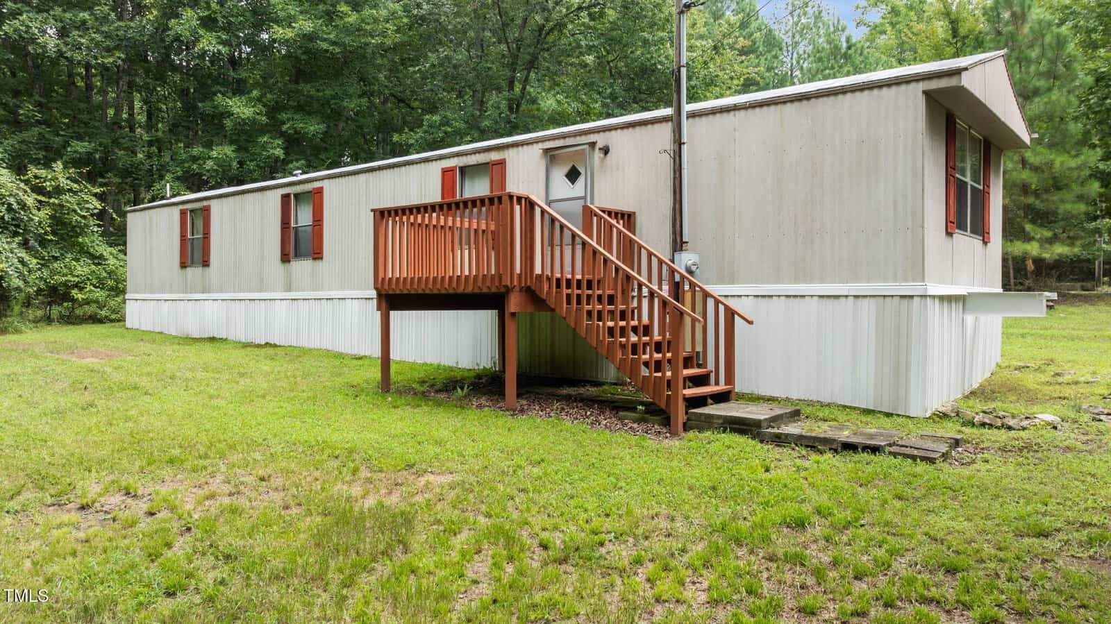 A single-wide beige mobile home with red shutters sits on a grassy lot. A wooden staircase with a railing leads to the front door. The home is surrounded by trees and has a white skirting around the base.