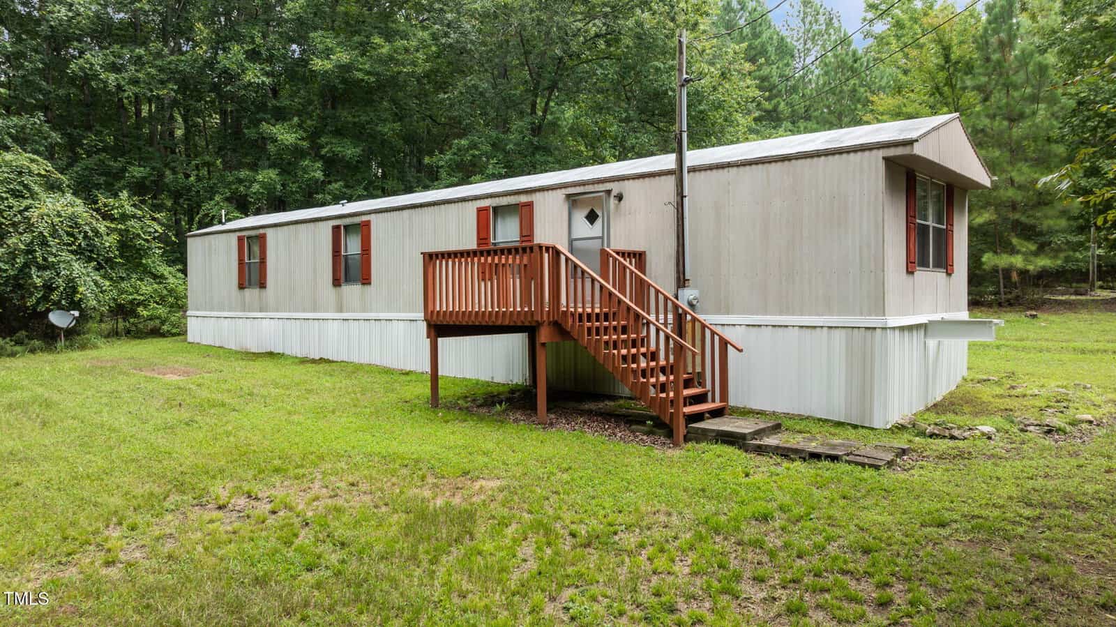 A single-wide mobile home with beige siding and red shutters, elevated wooden front steps, and a small porch, set on a grassy lot with trees in the background.