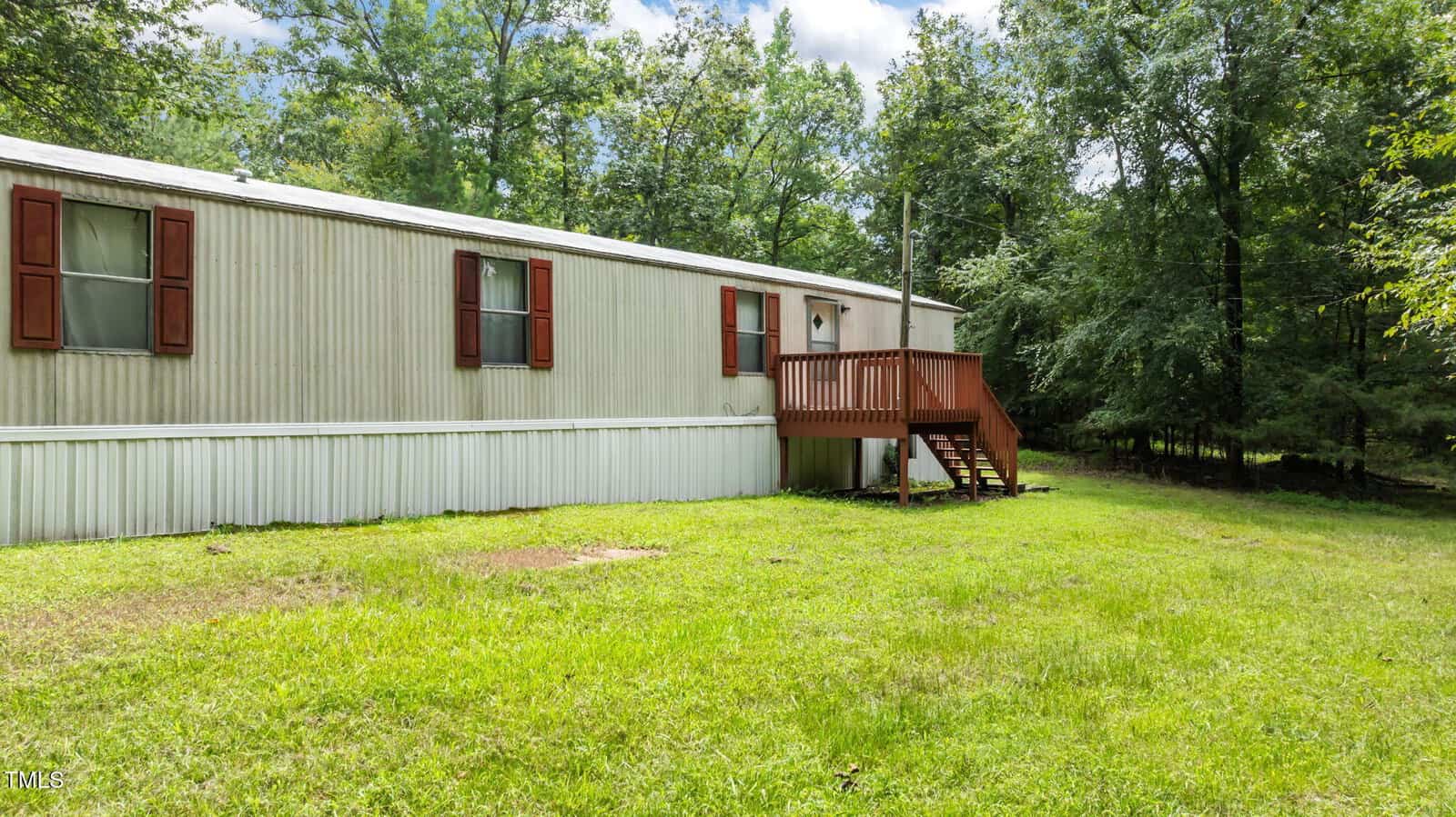 A beige mobile home with brown shutters sits on a grassy lot, surrounded by trees. It has a small wooden porch with steps leading up to the front door. The sky is partly cloudy above.