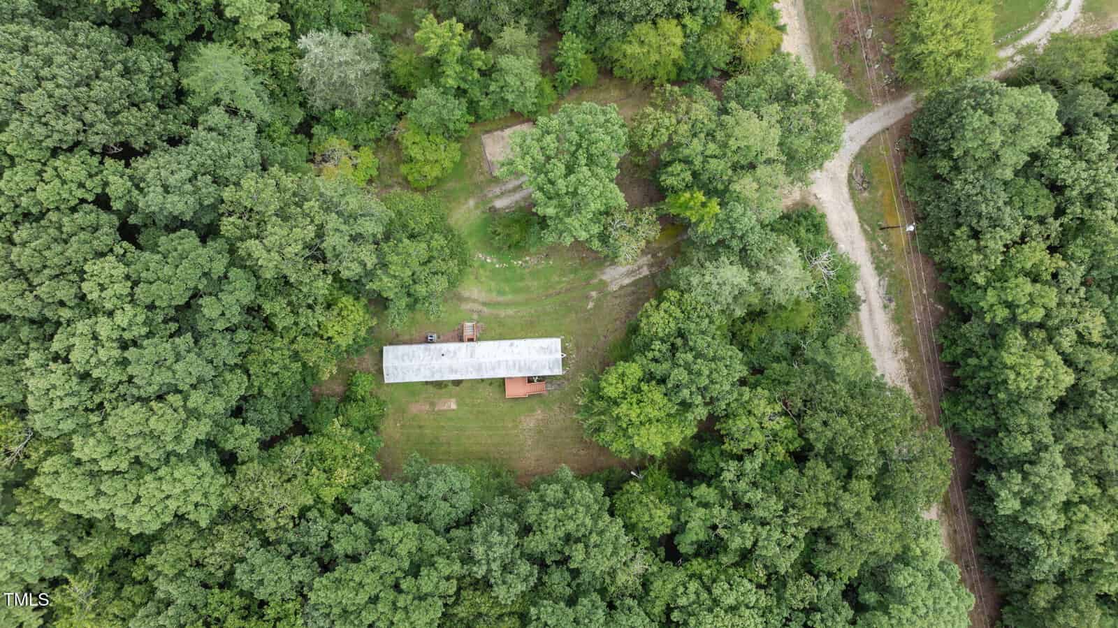 Aerial view of a rectangular building with a metal roof surrounded by dense green trees, a grassy clearing, and a dirt road curving nearby.