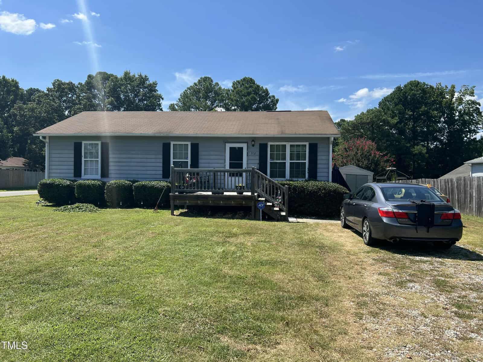 A single-story house with light blue siding, a front porch, and black shutters. A black sedan is parked on a gravel driveway, and the lawn is neatly mowed under a clear blue sky. Shrubs line the front of the house.