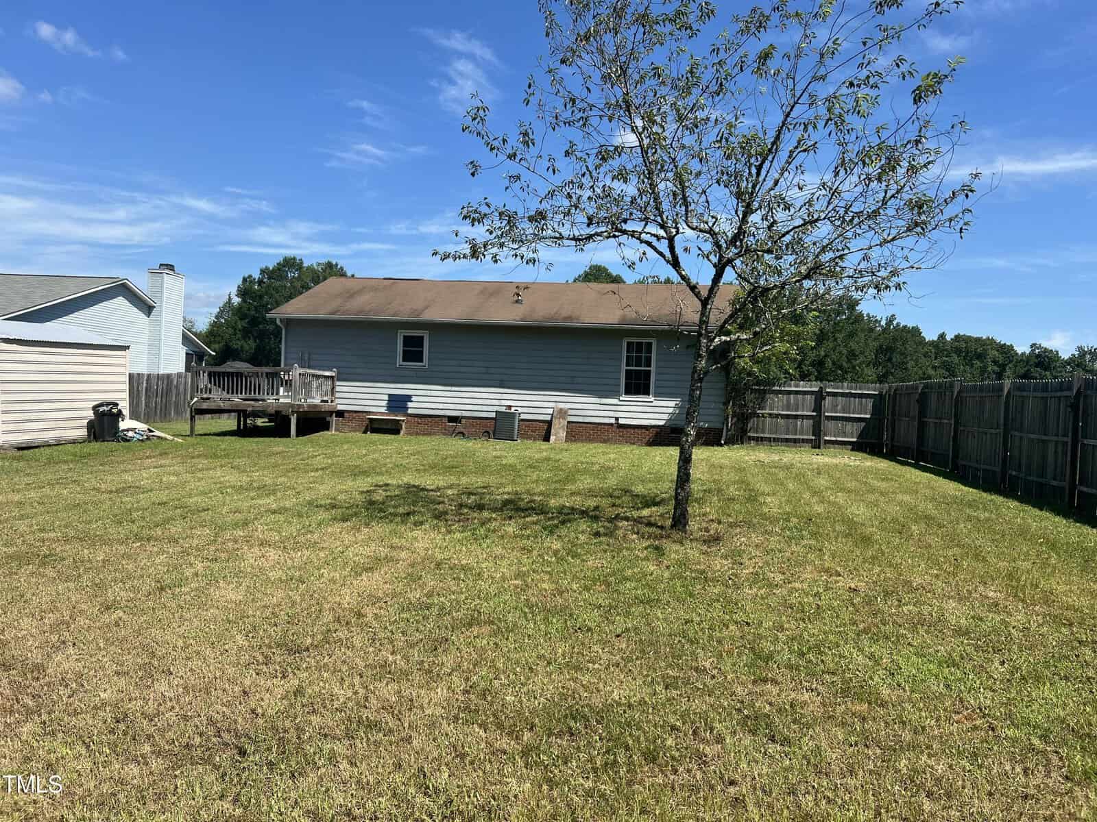 A backyard with a mowed lawn, a single tree, wooden fencing, and a light blue house with a deck and visible foundation. A clear blue sky is overhead.