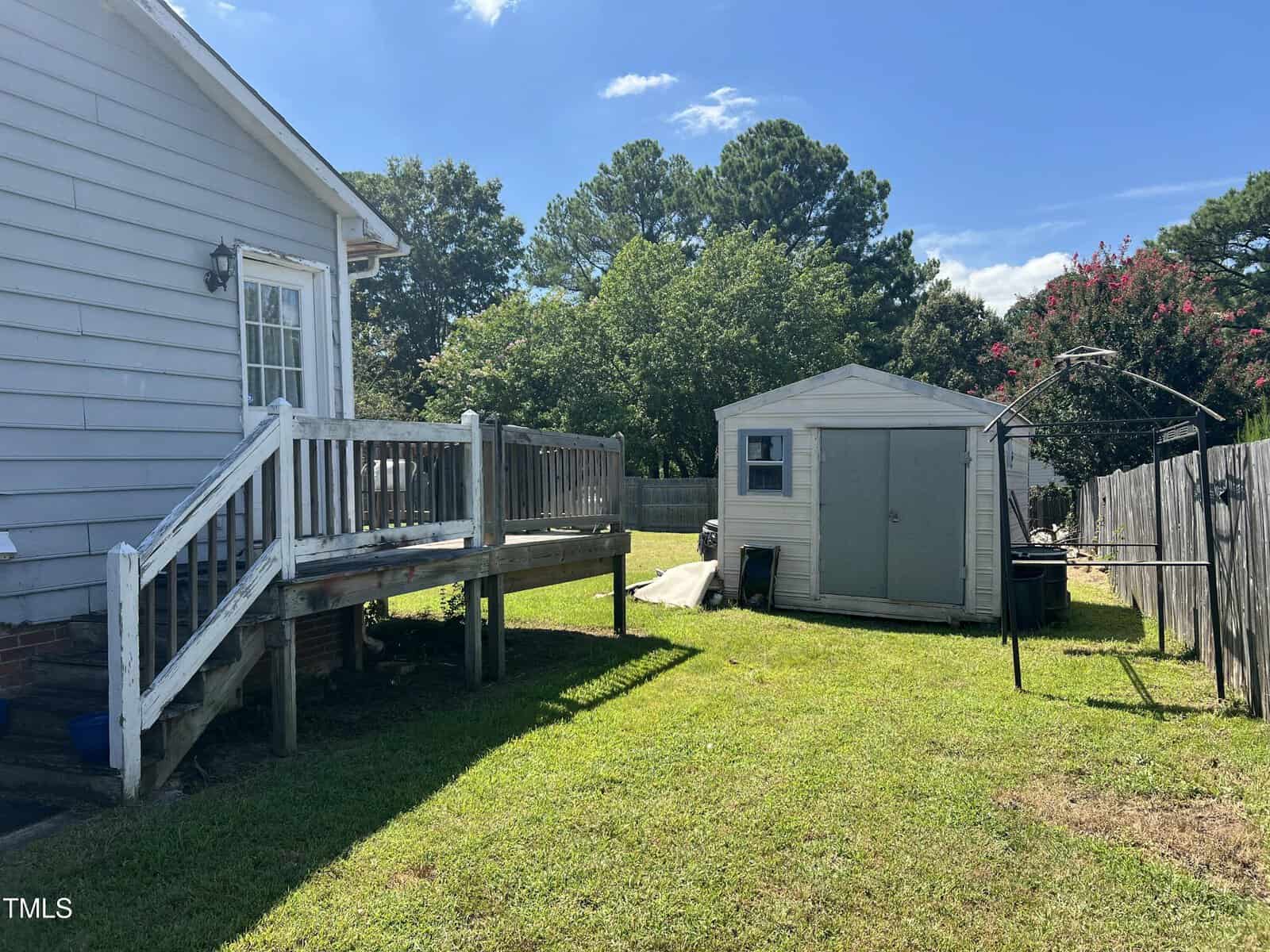 A small wooden deck with steps leads from a gray-sided house into a backyard with a grassy lawn, a white shed with a gray door, and a wooden fence under a sunny blue sky.