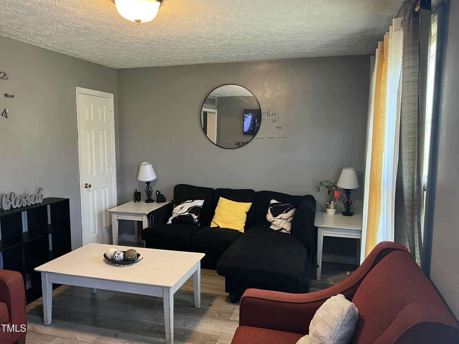 A living room with a black sectional sofa, yellow and white accent pillows, two side tables with lamps, a round wall mirror, a white coffee table, two red armchairs, and curtains covering windows on the right.