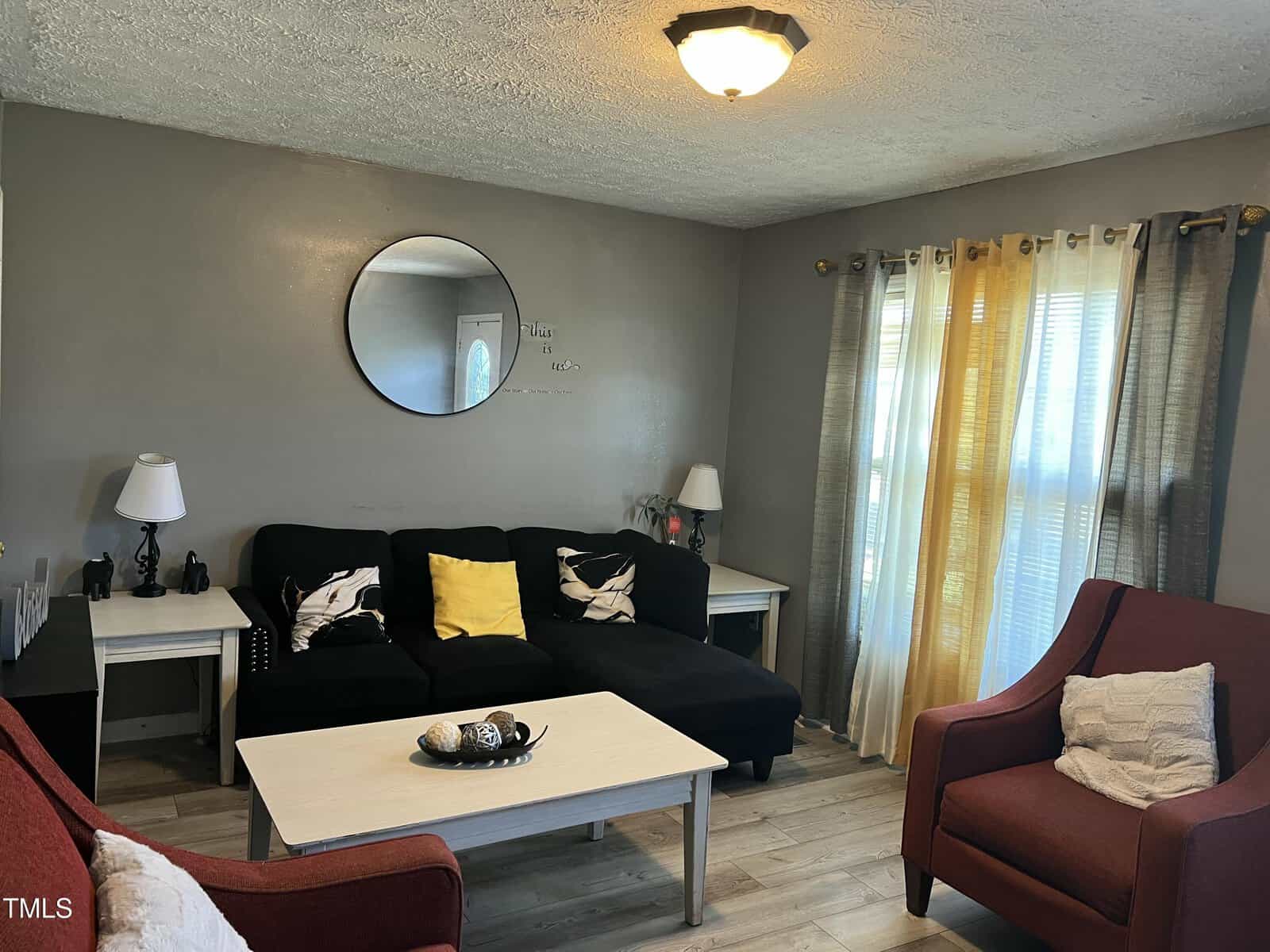 A small living room with gray walls, a black sectional sofa with patterned and yellow pillows, two red armchairs, a white coffee table, a round mirror on the wall, and sheer yellow curtains on the window.