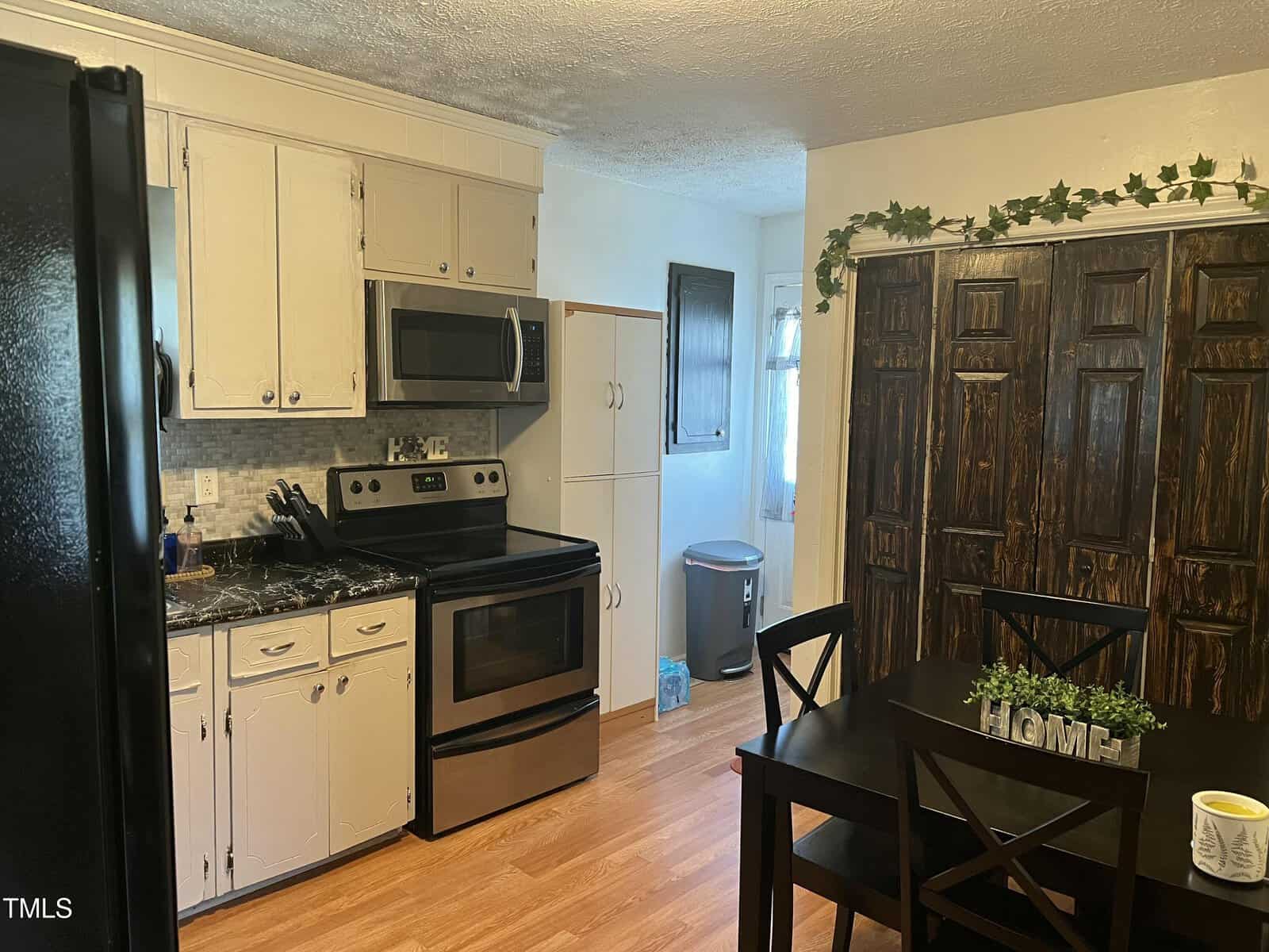 A kitchen with white cabinets, stainless steel appliances, a black refrigerator, a dining table with four chairs, and dark wooden closet doors. Decorative greenery hangs above the closet. The floor is light wood.