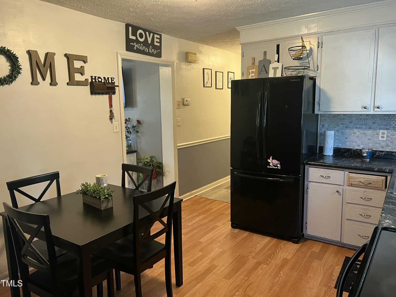 A cozy kitchen with a black fridge, white cabinets, black dining table with four chairs, and light wooden floor. “LOVE” and “HOME” wall decor are visible, along with framed pictures and a small wreath.