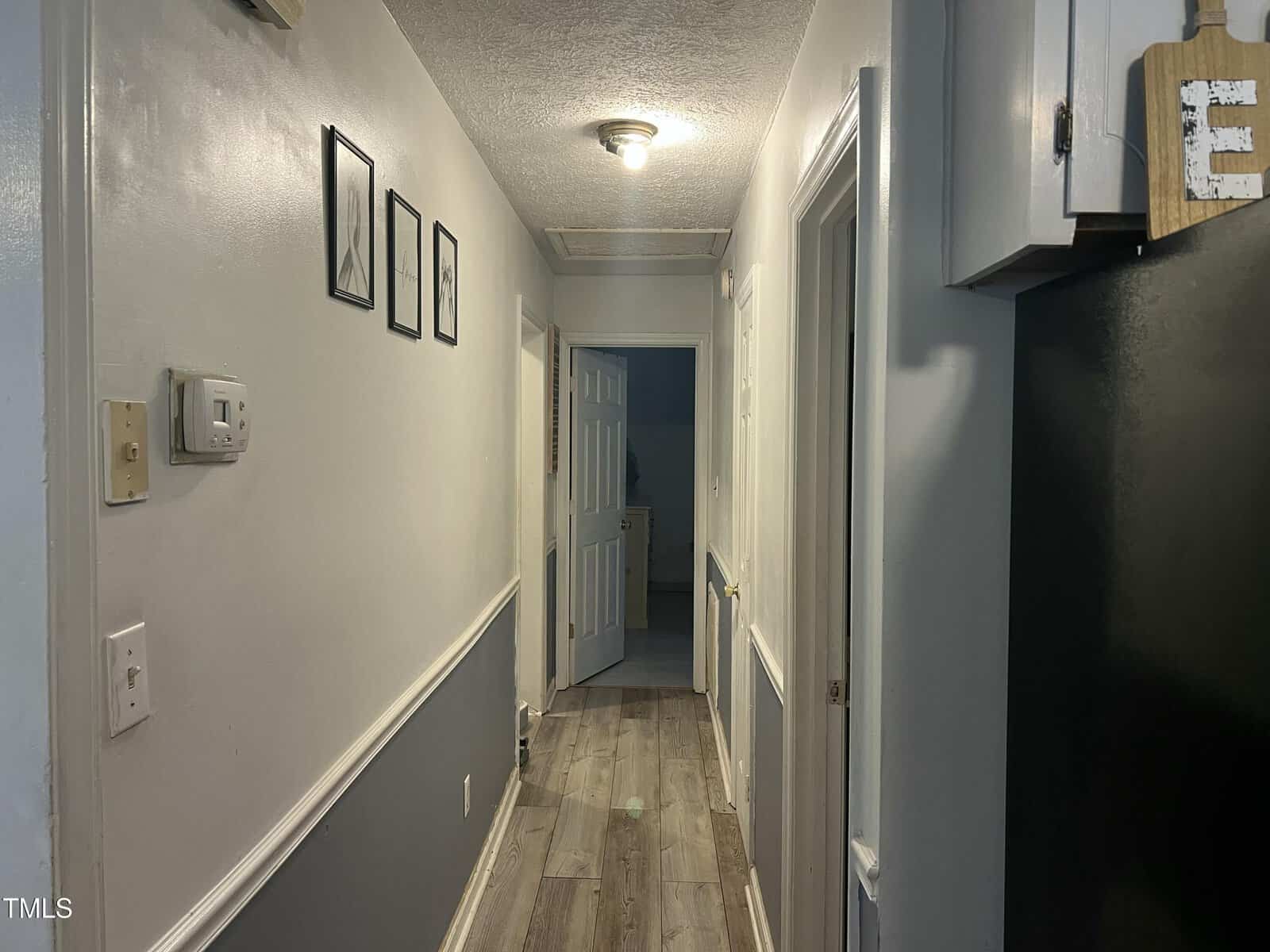 A hallway with gray walls, wood flooring, framed art on the left, a ceiling light, several doors, and a kitchen area partially visible on the right.