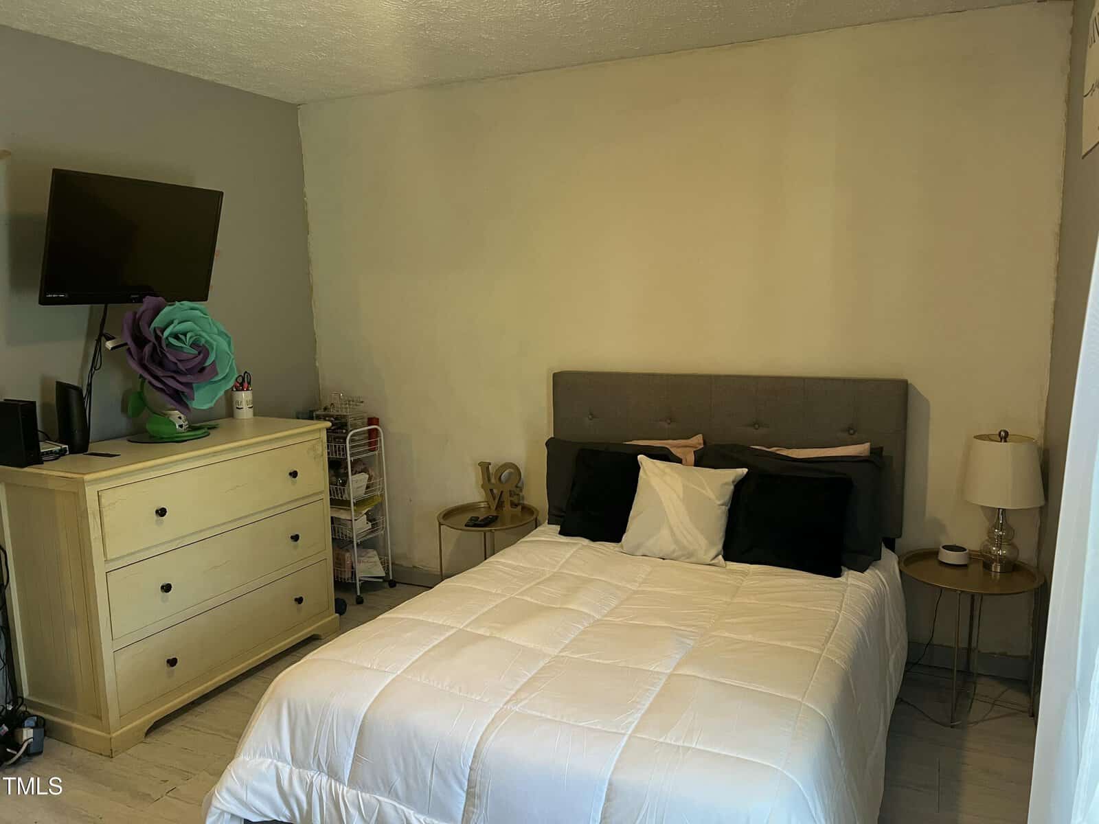 A neatly arranged bedroom with a bed, gray headboard, white comforter, and black pillows. A TV is mounted above a light dresser. A nightstand with a lamp and a floral decoration sit nearby.