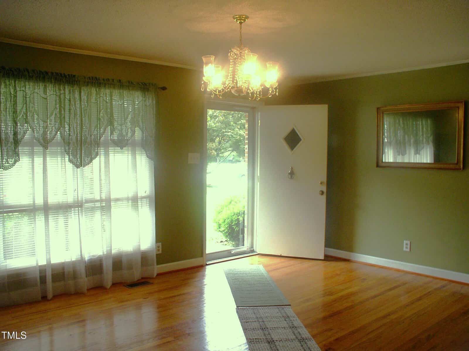 A room with hardwood floors, green walls, a window with sheer curtains, a wall mirror, and an open white door letting in natural light. A ceiling chandelier is turned on above a runner rug.