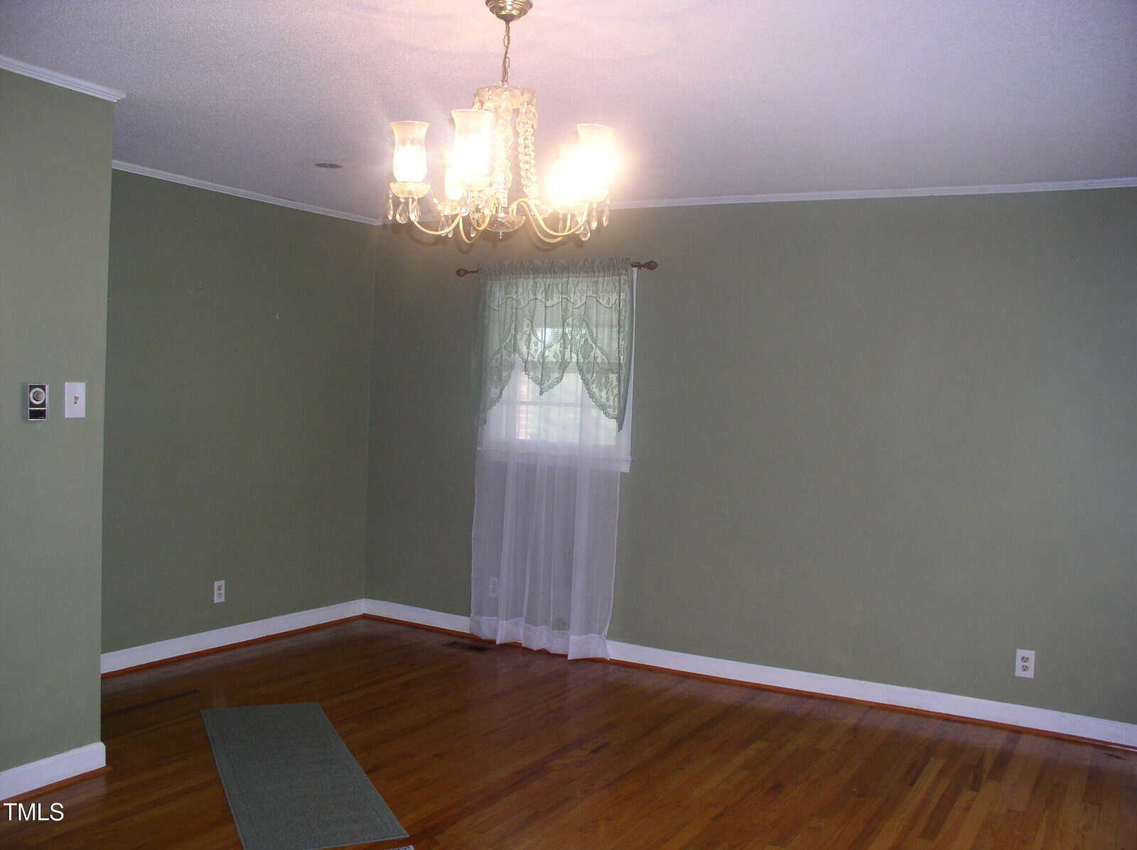 An empty room with green walls, a wooden floor, a window with sheer curtains, and a ceiling chandelier light fixture. A small floor mat is near one wall.