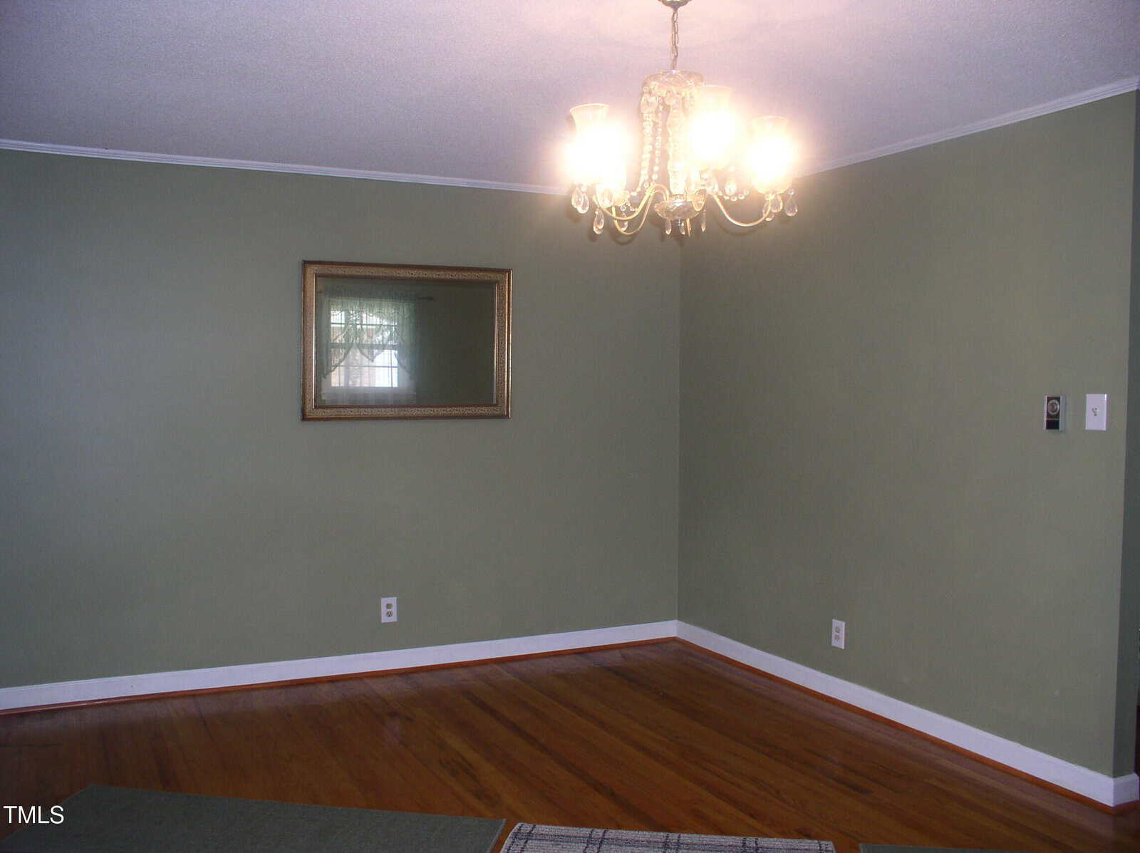 A mostly empty room with green walls, a wooden floor, a ceiling-mounted chandelier light, and a framed mirror on one wall. There is a light switch and an electrical outlet visible on the right wall.