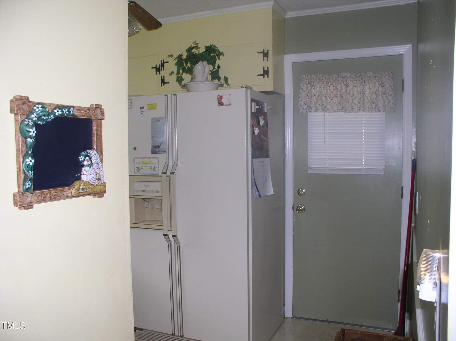 A kitchen corner with a white refrigerator, a green door with a small window and floral curtain, a framed chalkboard on a yellow wall, and a potted plant on top of the fridge.